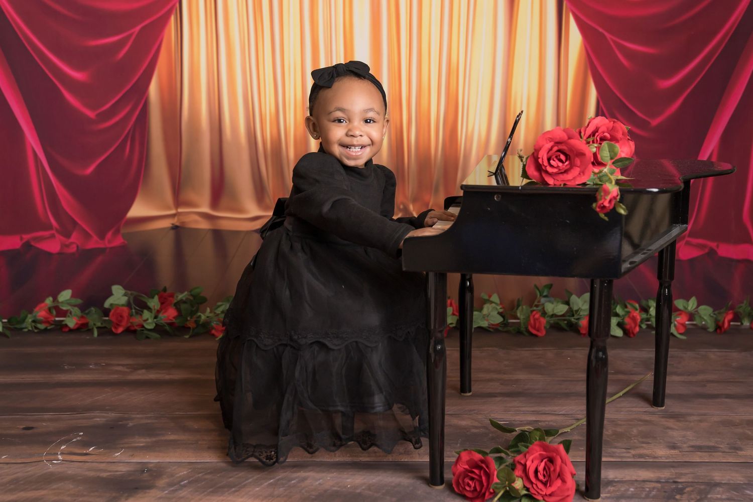 Child in black dress smiling while seated at piano with red roses and curtain backdrop.