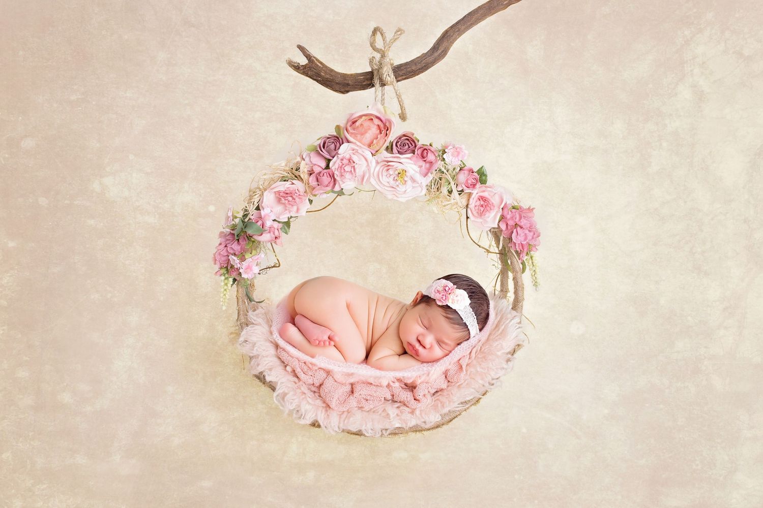 Newborn baby sleeping peacefully in a floral wreath basket suspended from a branch in a soft pink nursery photo.