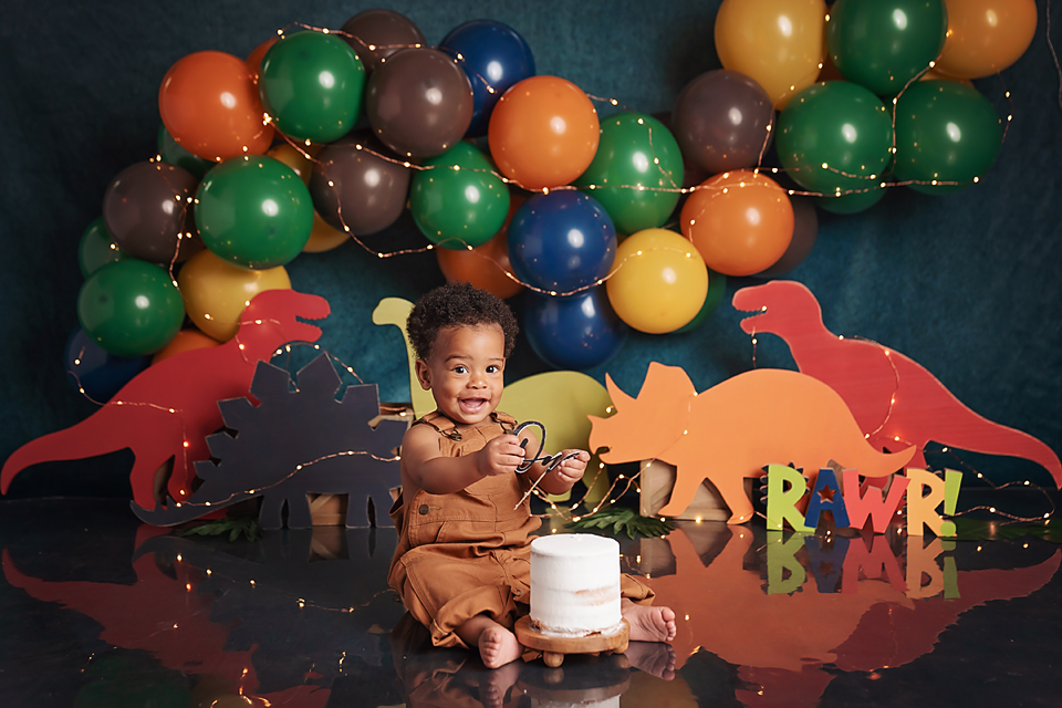 Baby's first birthday cake smash with dinosaur theme, colorful balloons, and RAWR letters on dark teal backdrop.