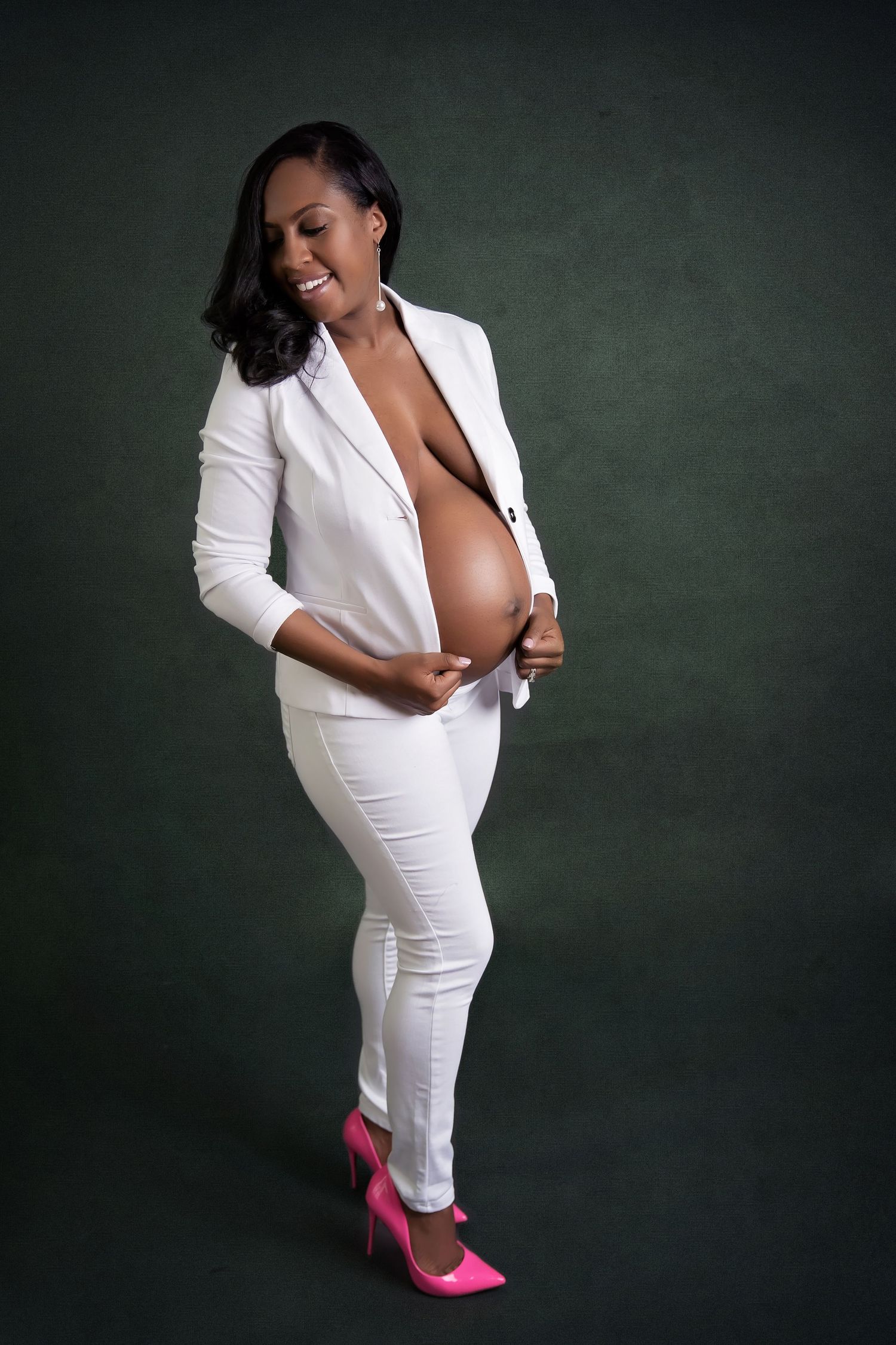 Stylish maternity photo in white suit and pink heels against dark green background.