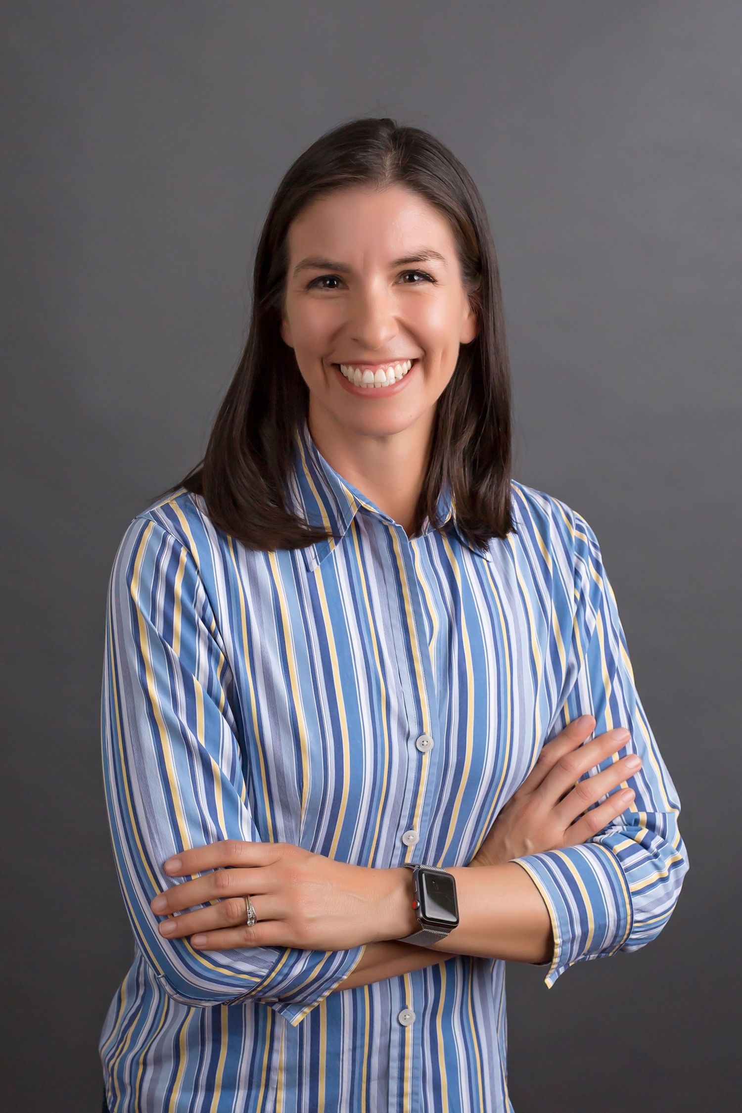 Woman in blue striped shirt posing with crossed arms for professional headshot against gray backdrop
