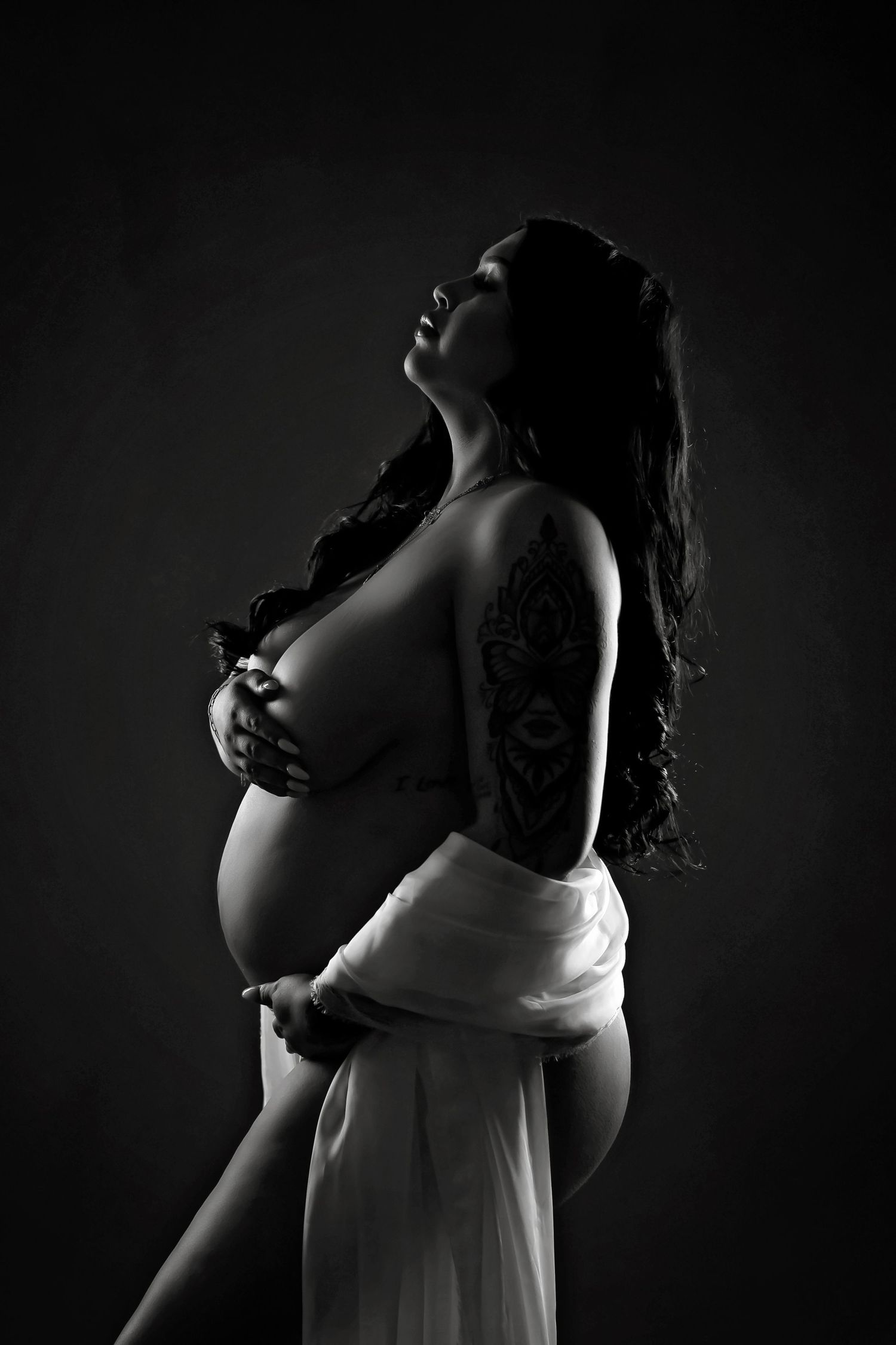 Black and white silhouette of a pregnant woman with tattoos posing gracefully in soft white fabric against dark background.