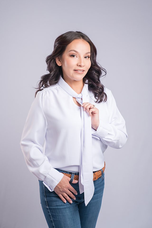 A young woman in a white blouse and jeans poses confidently against a gray background, holding her collar.