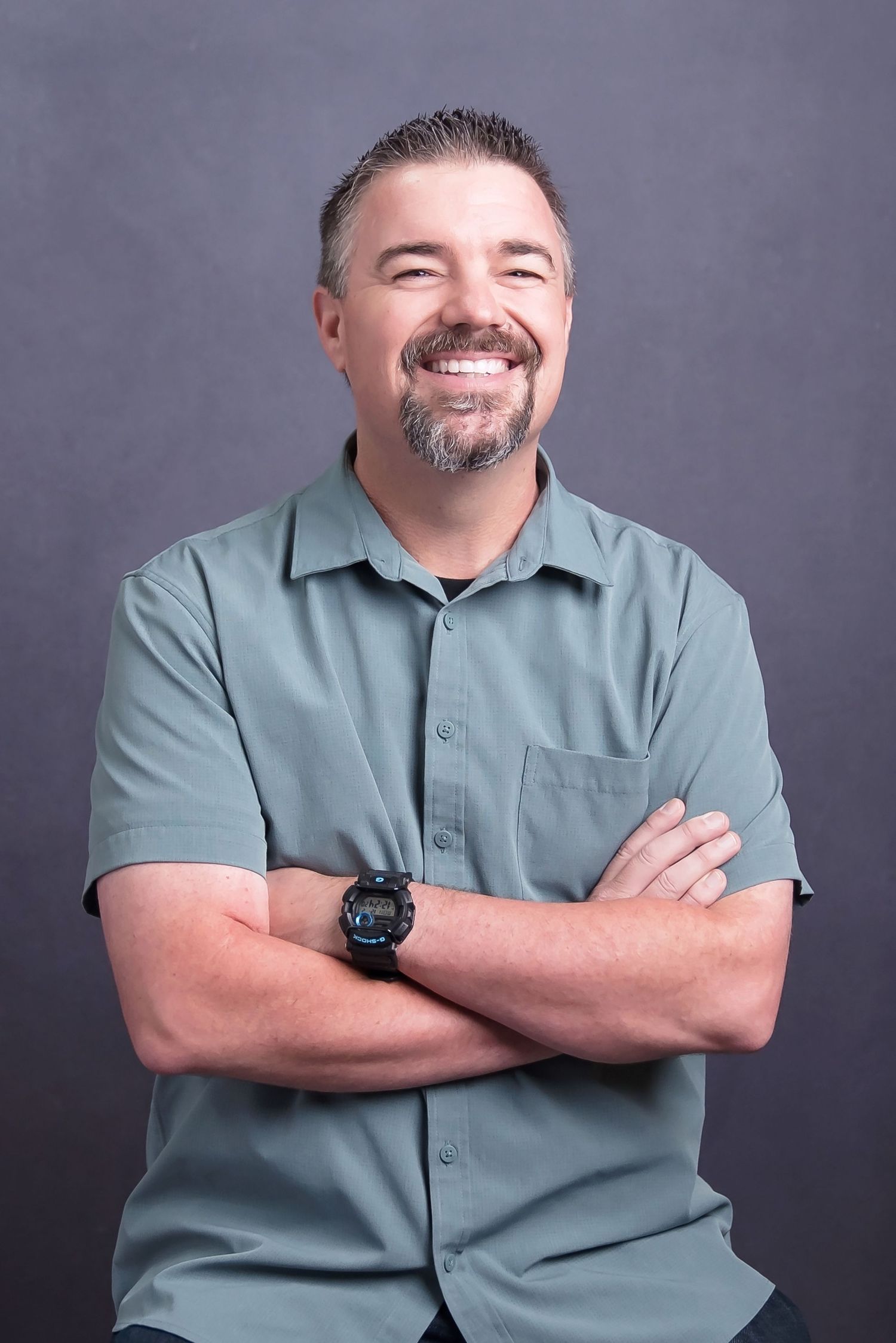 Professional portrait of man in sage green polo shirt posing with crossed arms against dark background