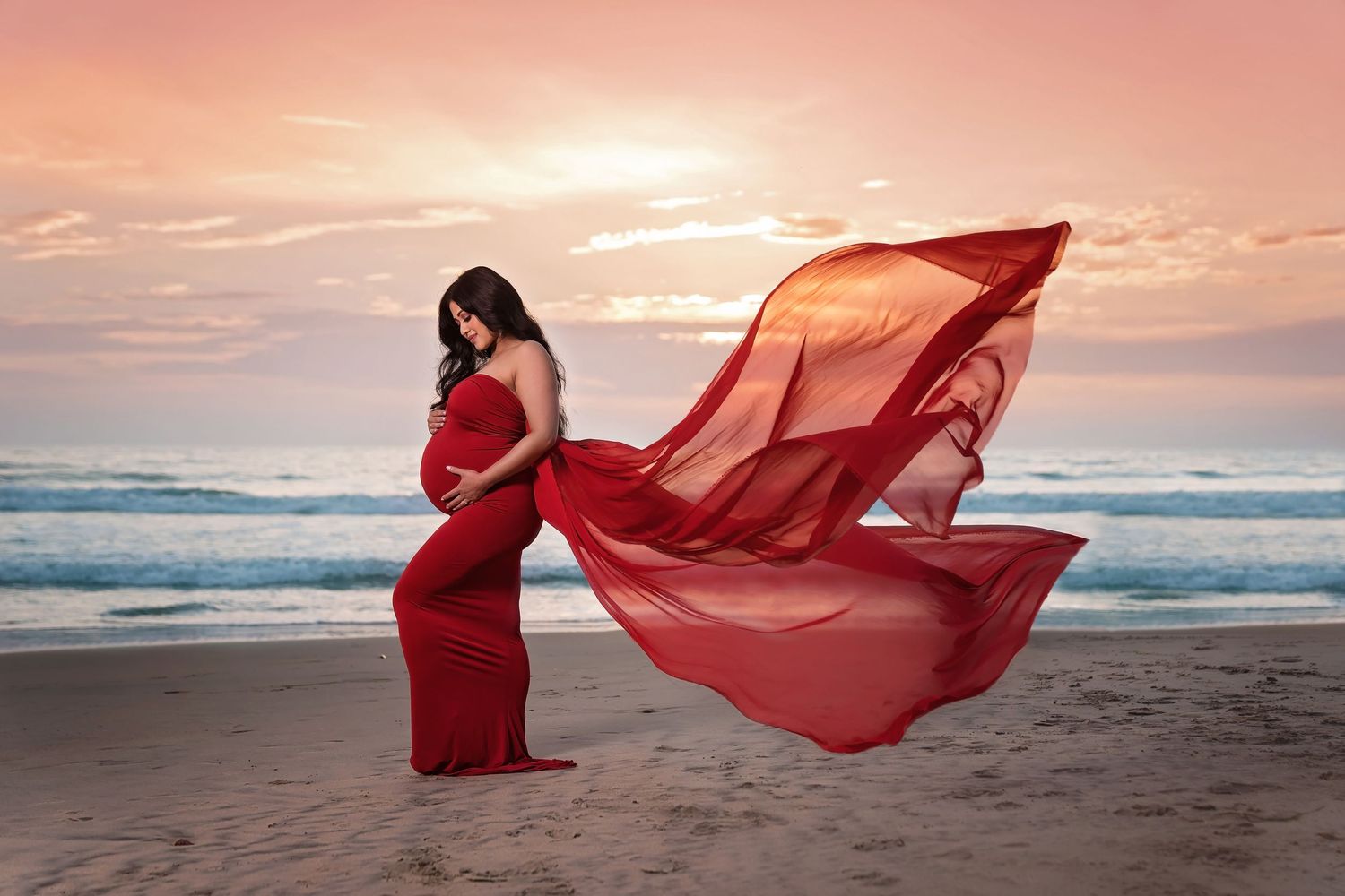 Maternity beach photo with flowing red dress catching wind during golden sunset. San Diego Maternity Photographer
