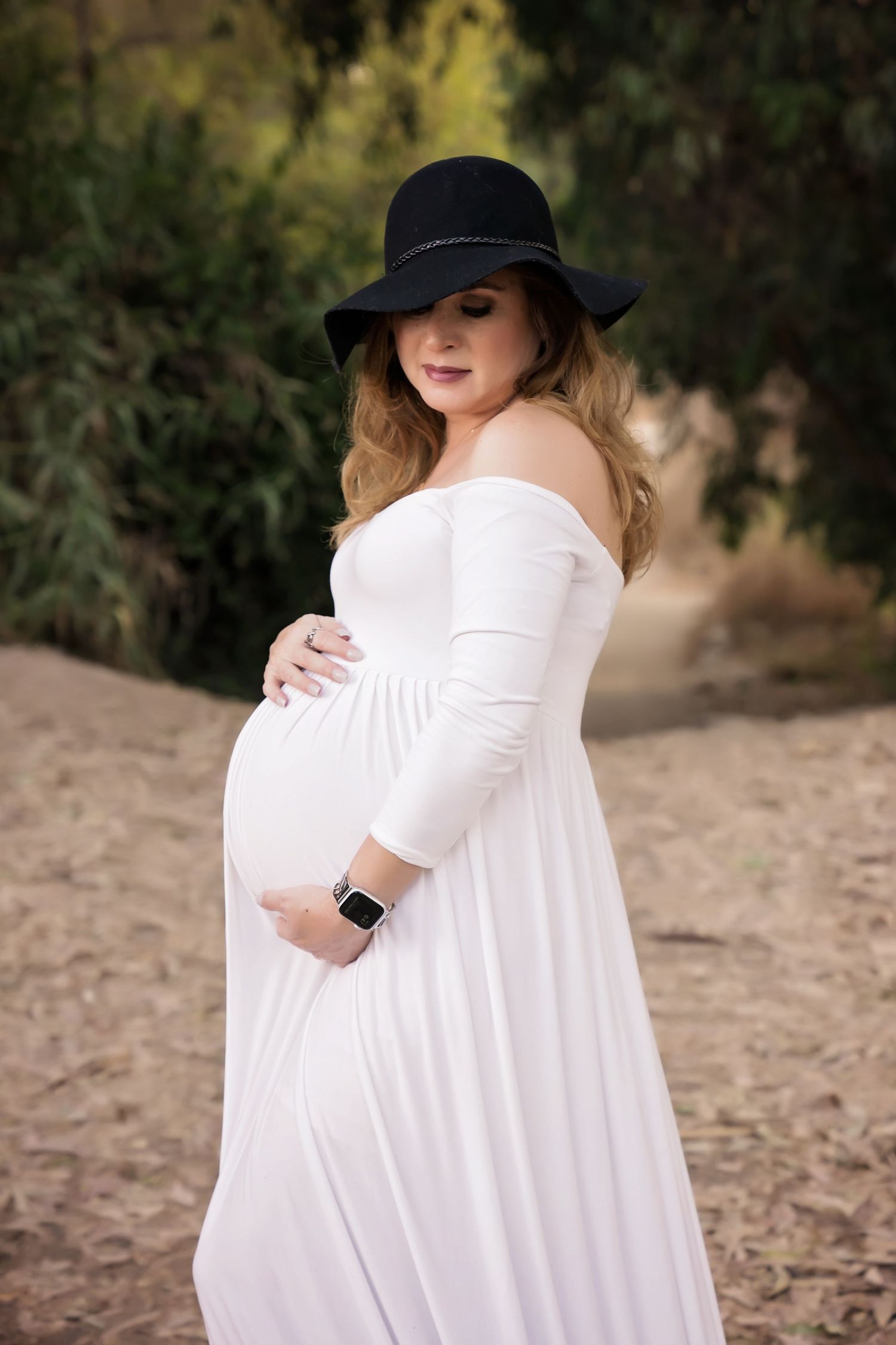 Elegant maternity photoshoot featuring a white dress and wide-brimmed black hat in a natural setting. San Diego Maternity Photographer