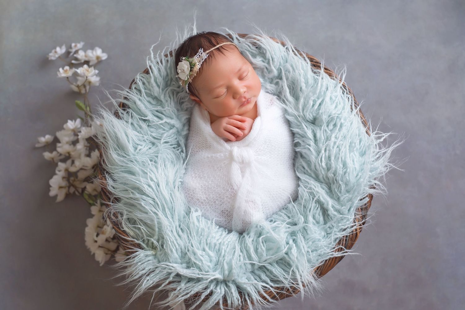 Newborn sleeping in round basket with soft mint green fur and white flowers. San Diego Newborn Photographer