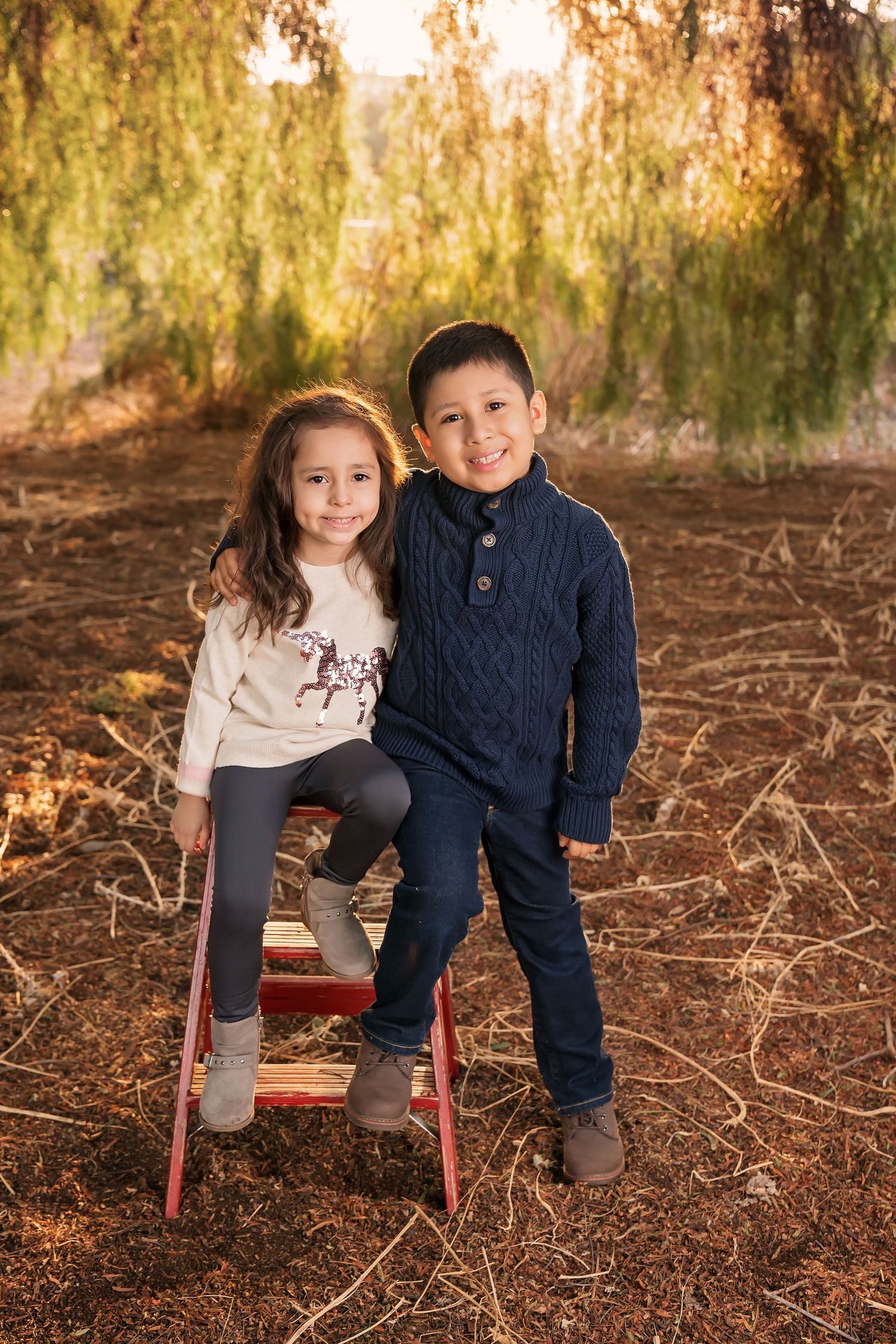 Two children seated together on a red stool under willow trees in a peaceful outdoor scene