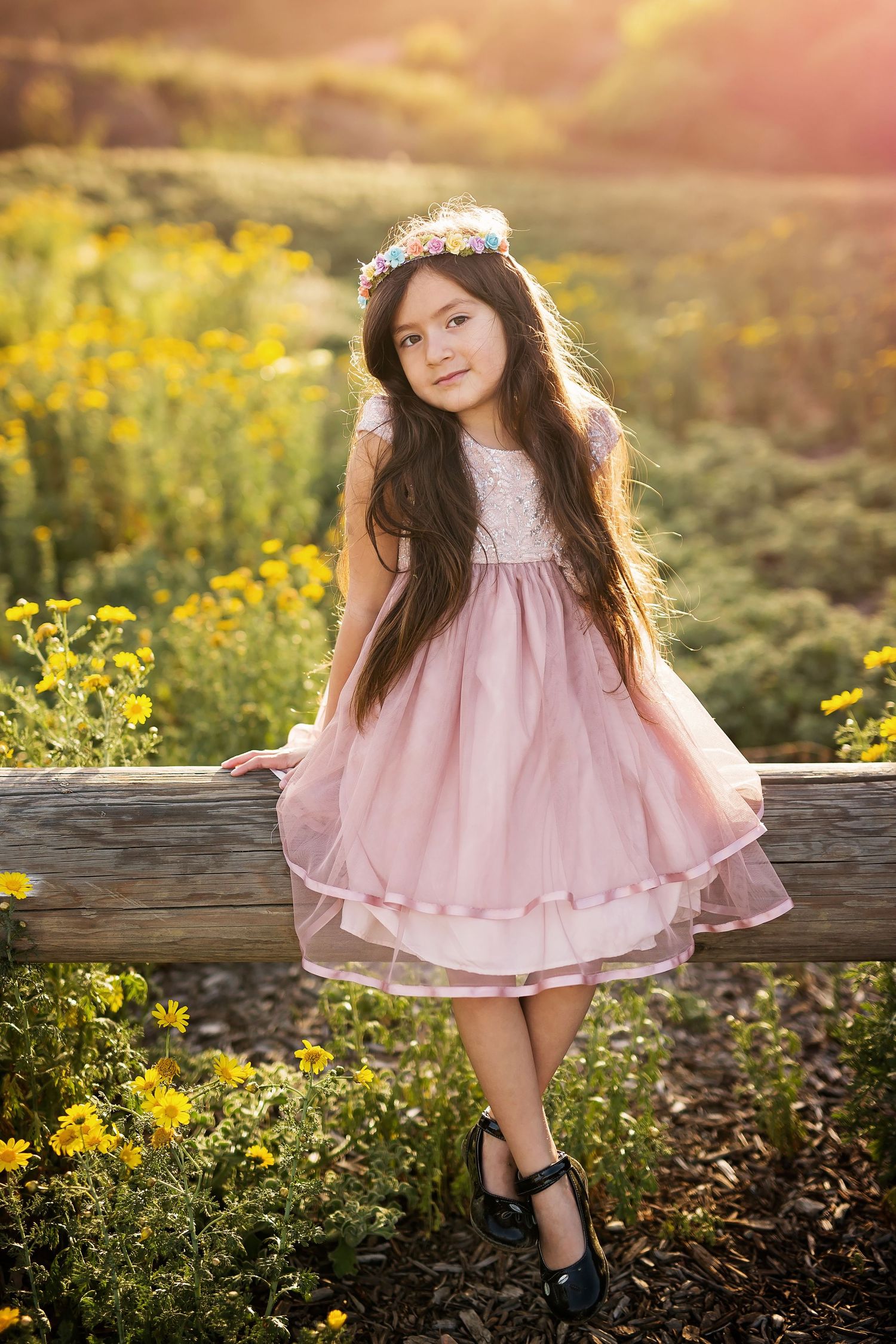 Child in a pink dress sitting on a rustic wooden fence among yellow wildflowers at sunset