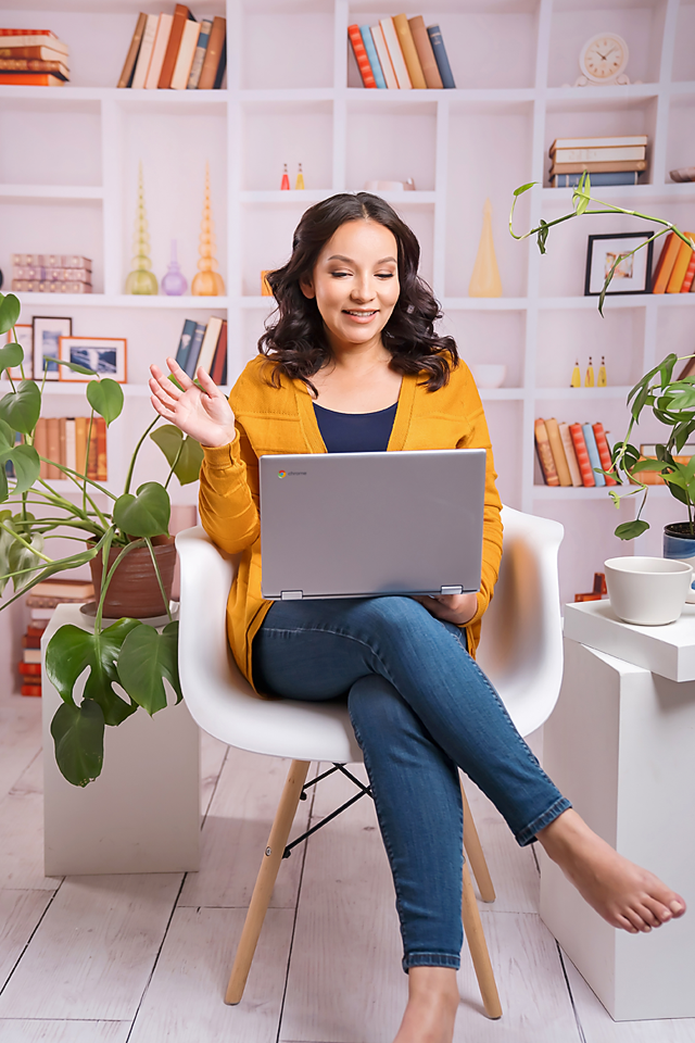 Woman in yellow cardigan waving during video call on laptop while sitting in modern white chair with bookshelves behind.