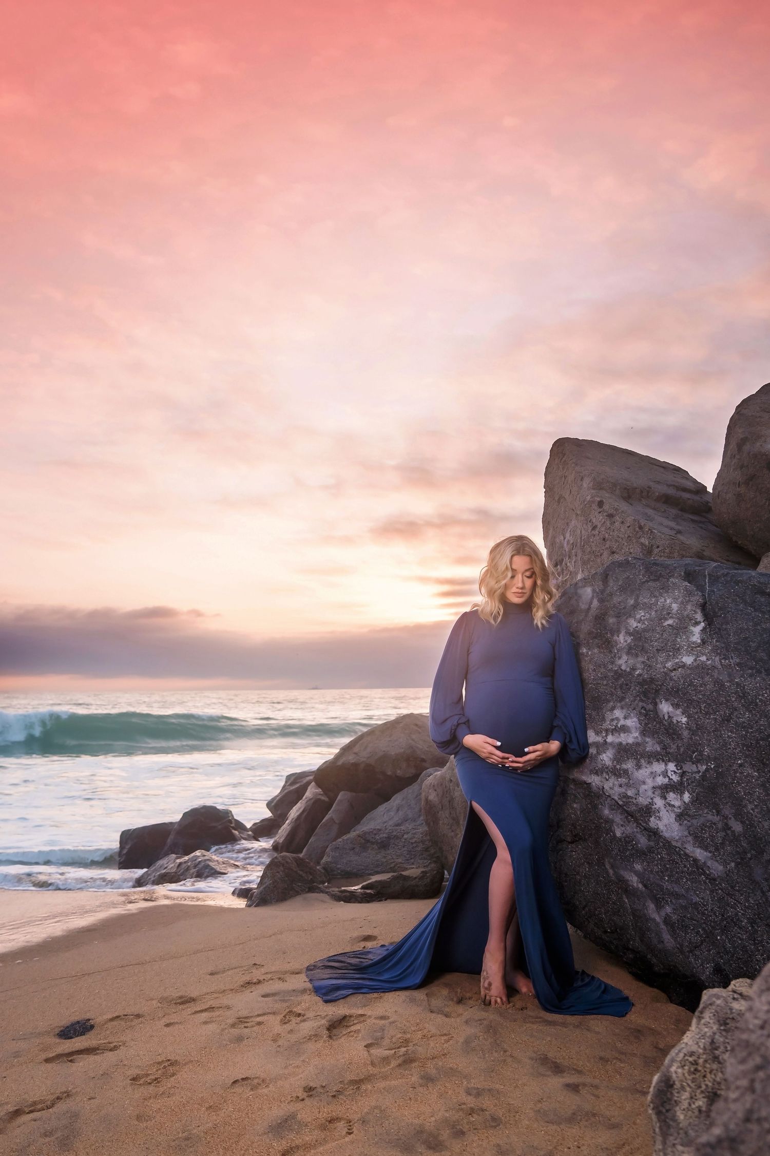 expectant mother posing against large rocks on beach during golden hour with crashing waves in san diego.