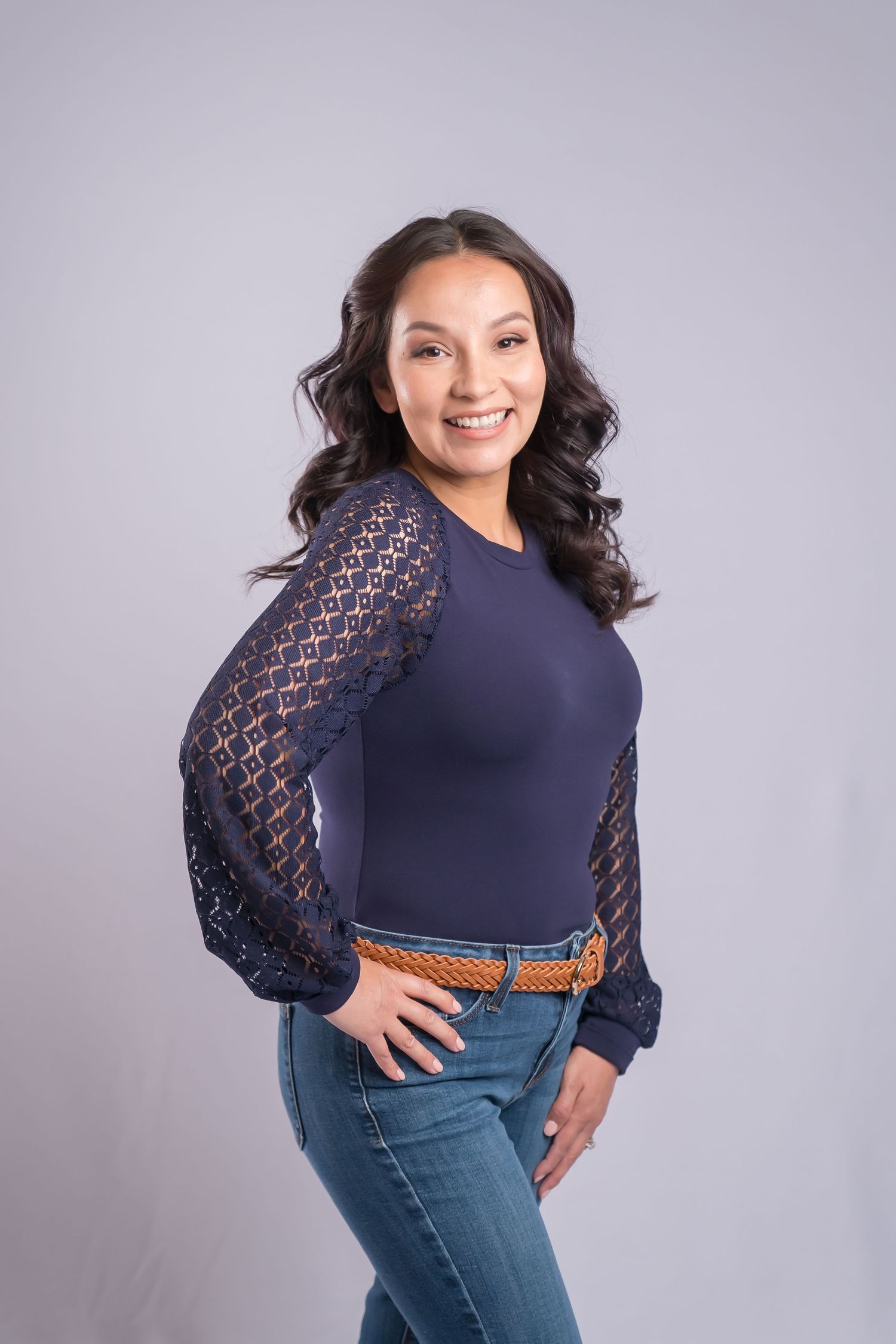 A person in a navy lace-sleeve top and jeans poses confidently against a gray background. Professional san diego Heashot