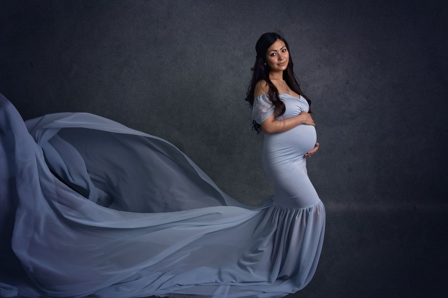 Elegant maternity photo with flowing blue dress fabric against dark backdrop.