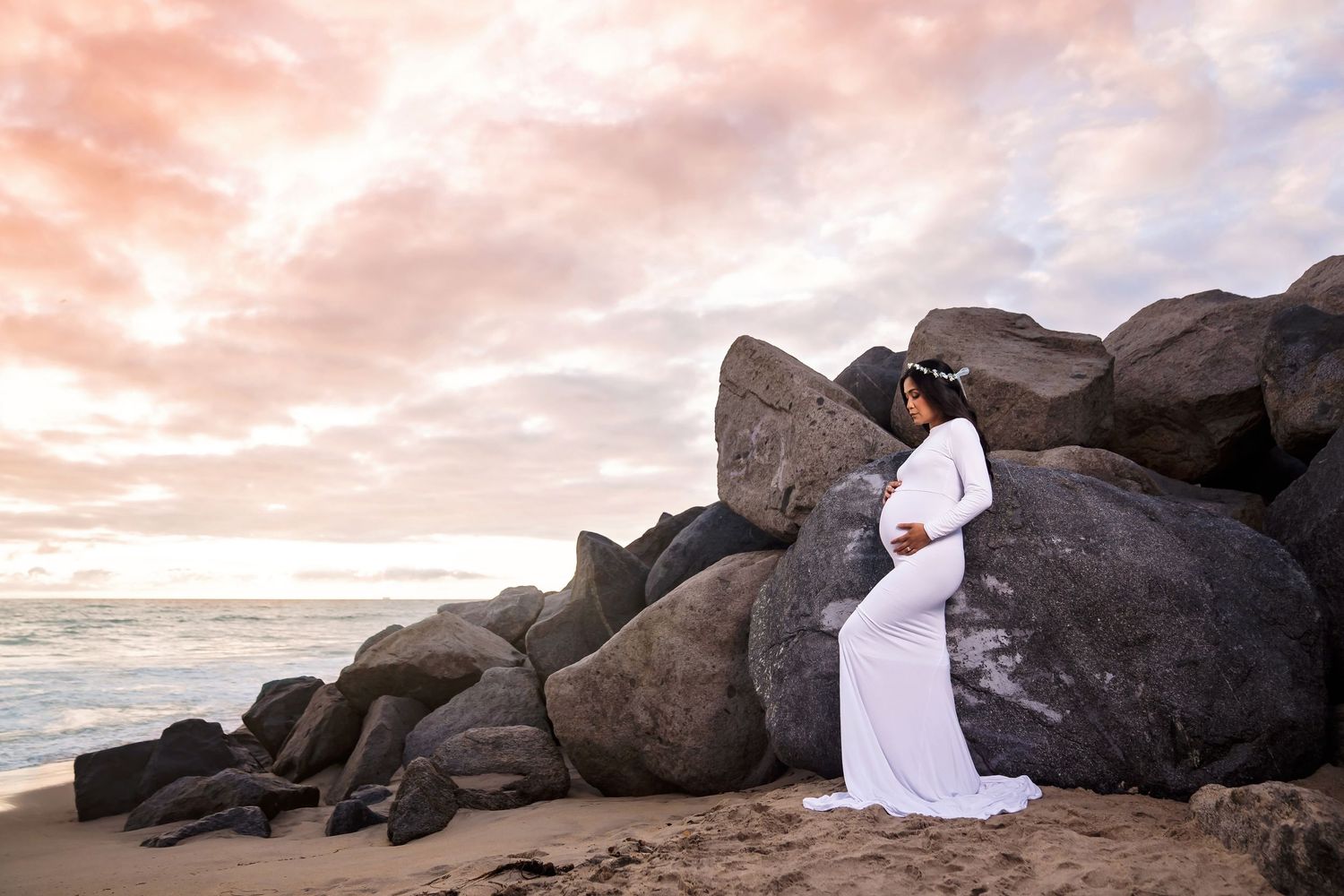 Dreamy beach photoshoot with figures posed among dark rocks at sunset. San Diego Maternity Photographer