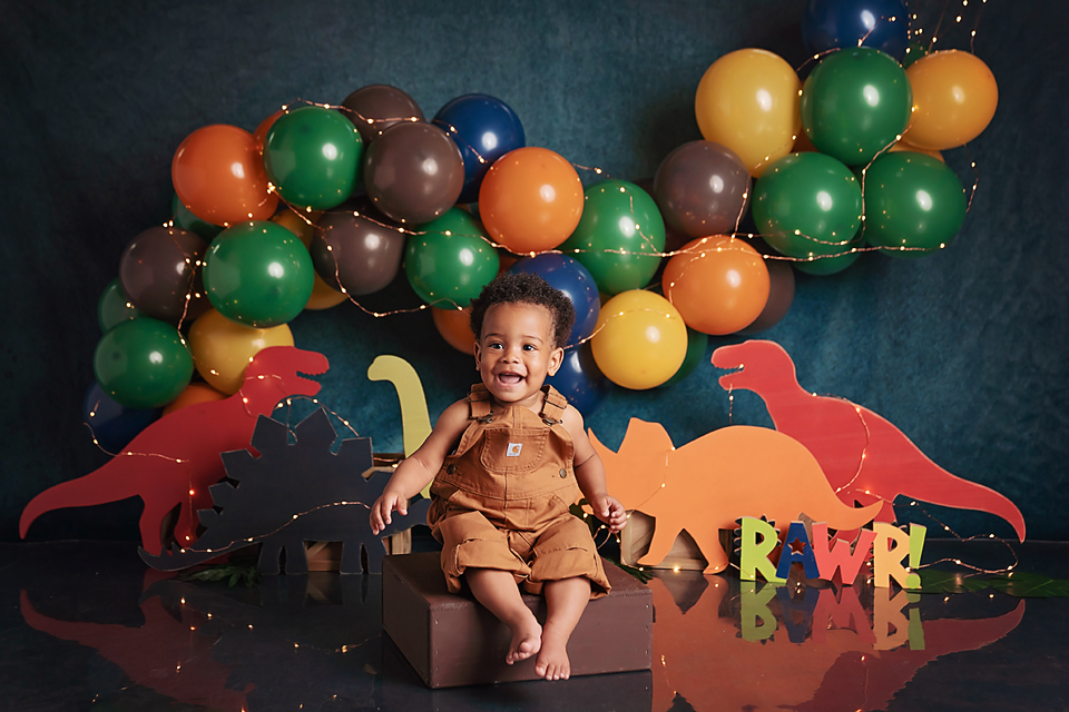 Baby's first birthday dinosaur smash cake session with colorful balloons and dino cutouts spelling RAWR.