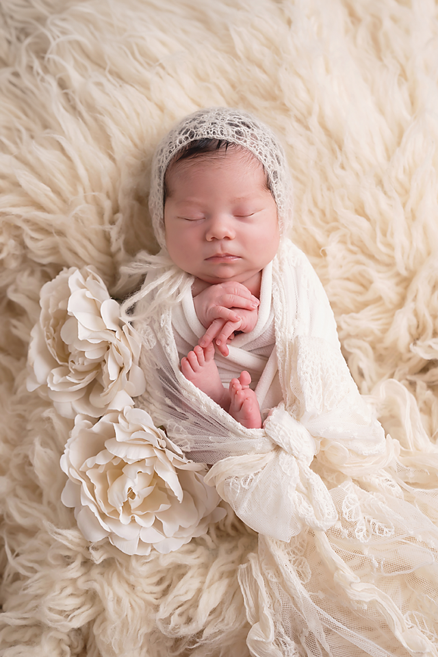Sleeping newborn baby wrapped in cream lace swaddle with floral accents and knit bonnet on fluffy white fur backdrop.