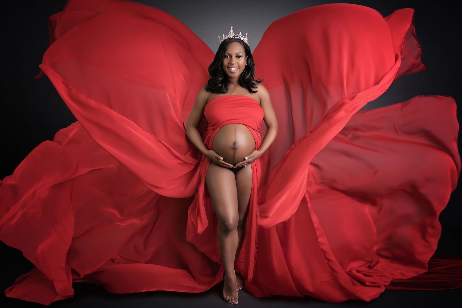 Dramatic maternity photo with flowing red fabric against black background.
