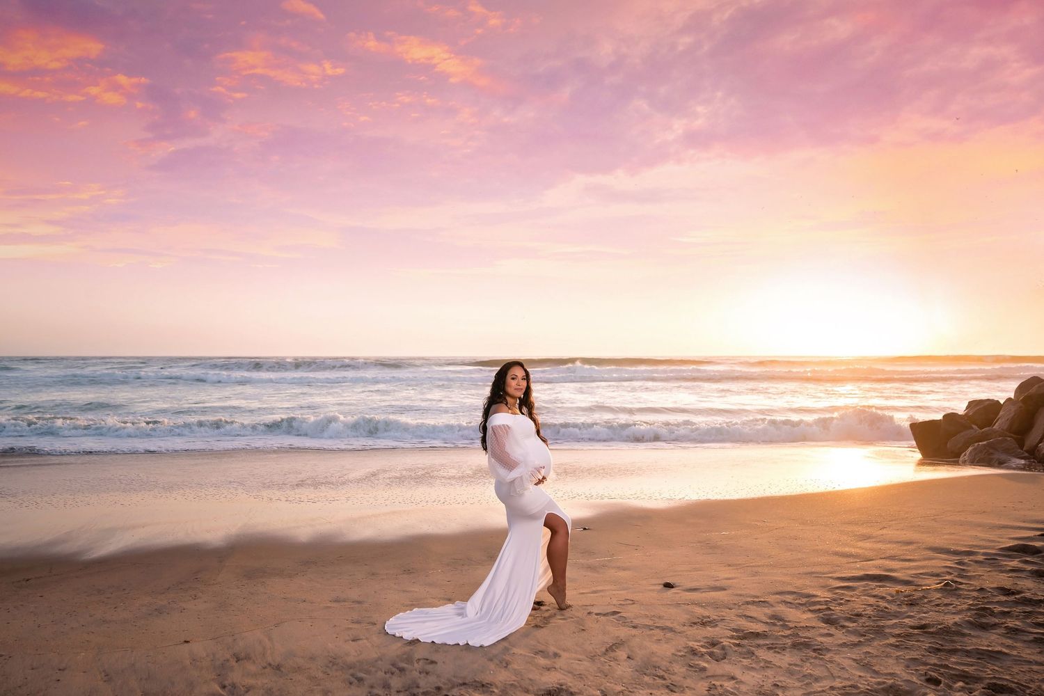 a pregnant woman wearing a white flowing dress catches the ocean breeze during a golden hour beach photoshoot. San Diego Maternity Photograp