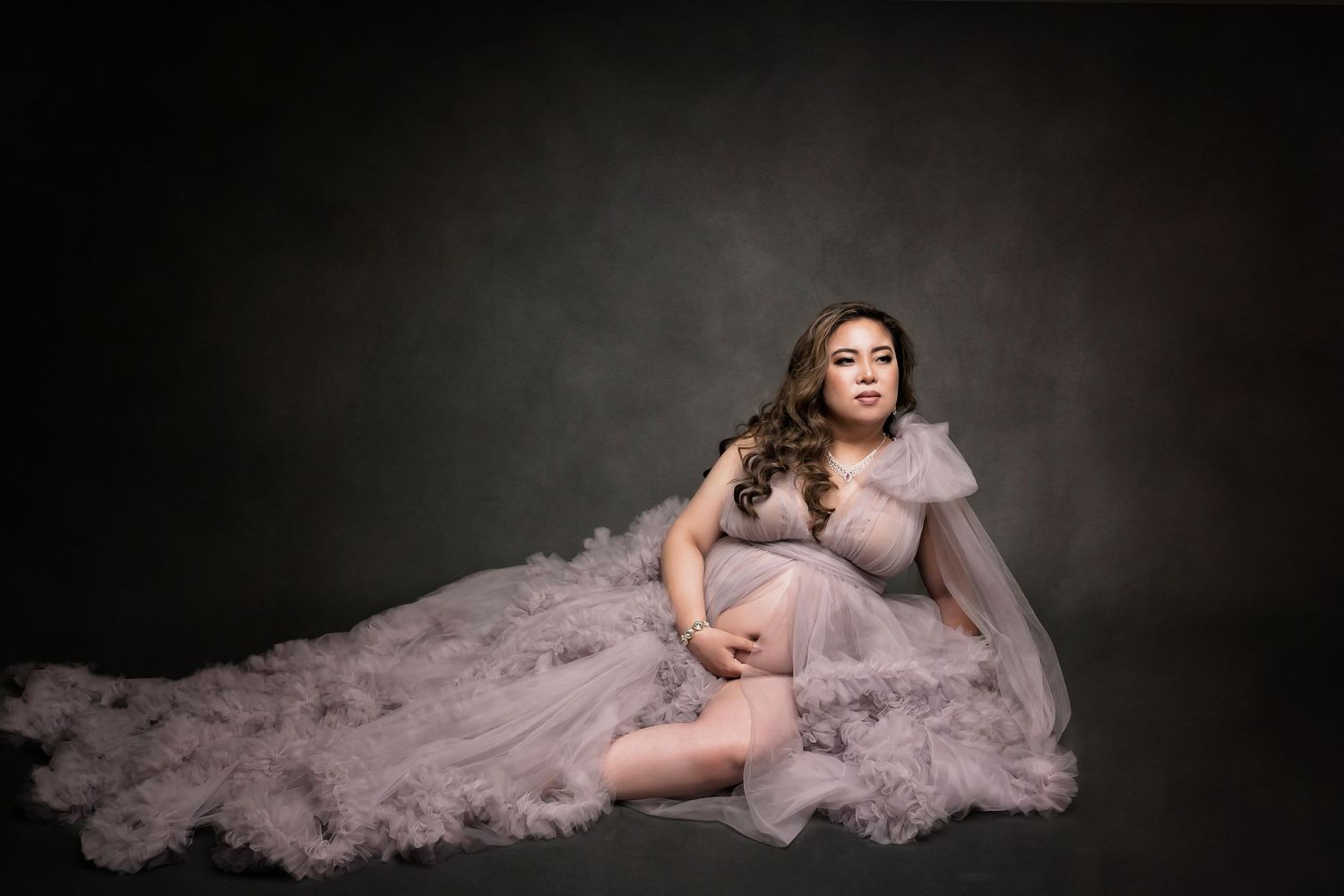 Pregnant woman in flowing lavender tulle gown poses gracefully on studio floor against dark backdrop.