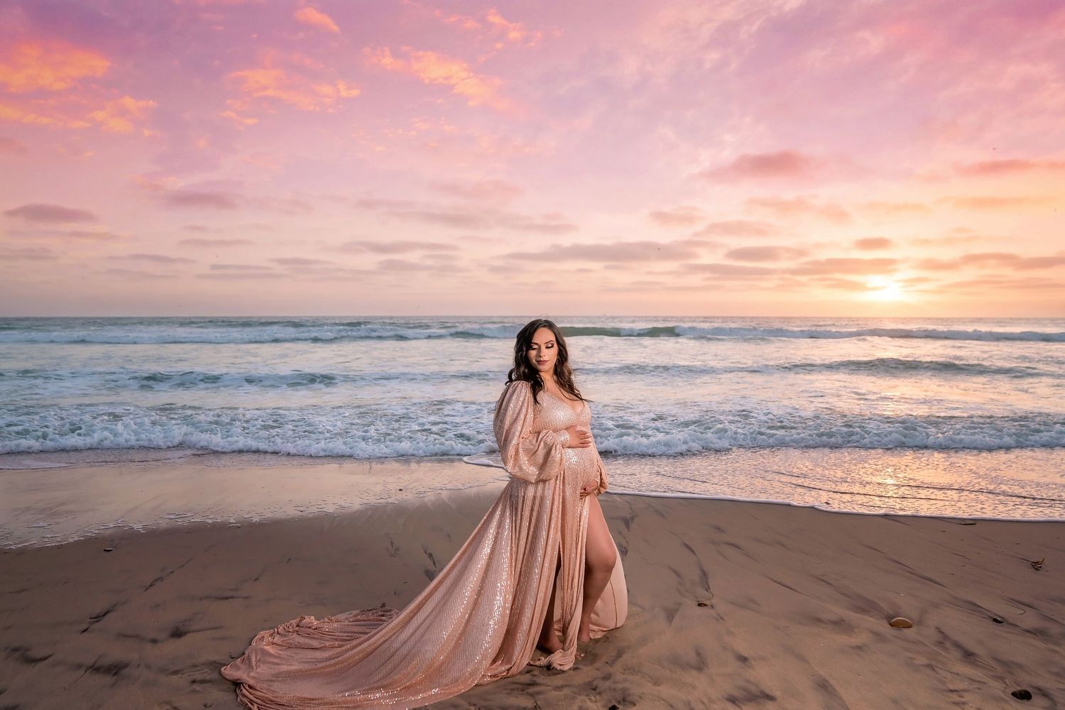 Romantic maternity beach photoshoot at sunset with flowing pink gown against ocean waves and sandy shore. San Diego Maternity Photographer