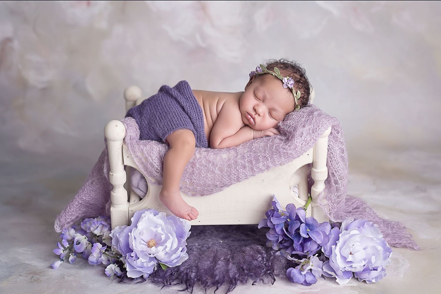 Newborn resting on white bench with purple floral decorations in soft lighting. San Diego Newborn Photographer