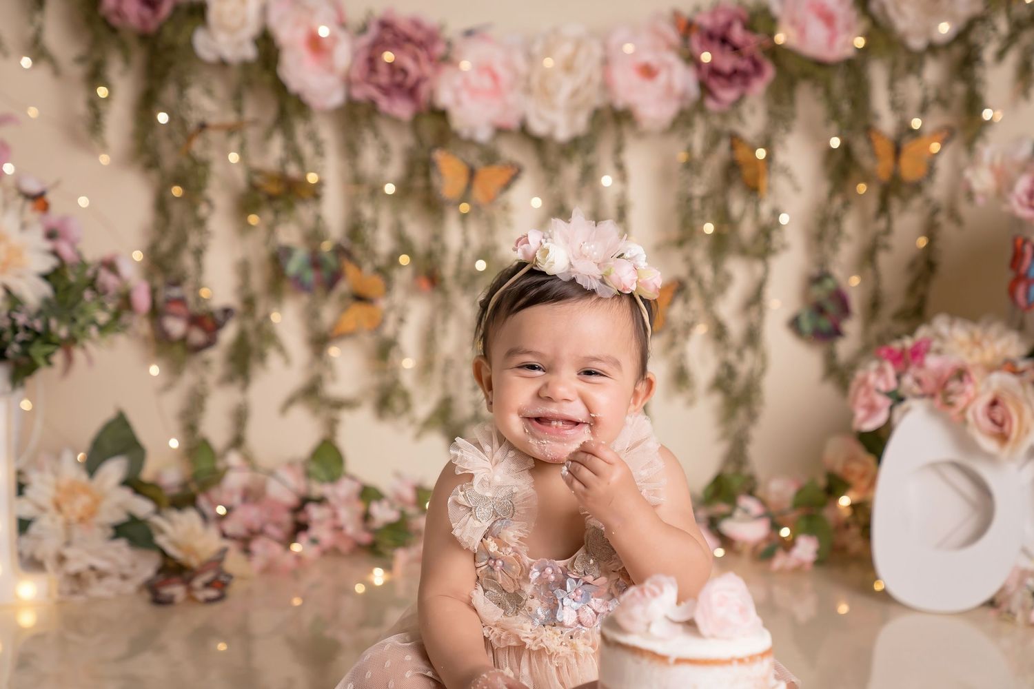 smash cake session with smiling baby girl in pink lace dress with floral crown sits before a flower and butterfly decorated backdrop.