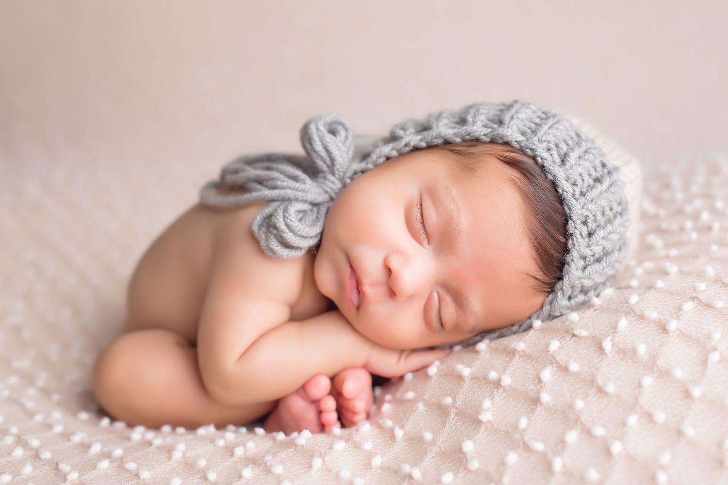 Sleeping newborn wearing gray knit bonnet posed on soft textured backdrop. San Diego Newborn Photographer