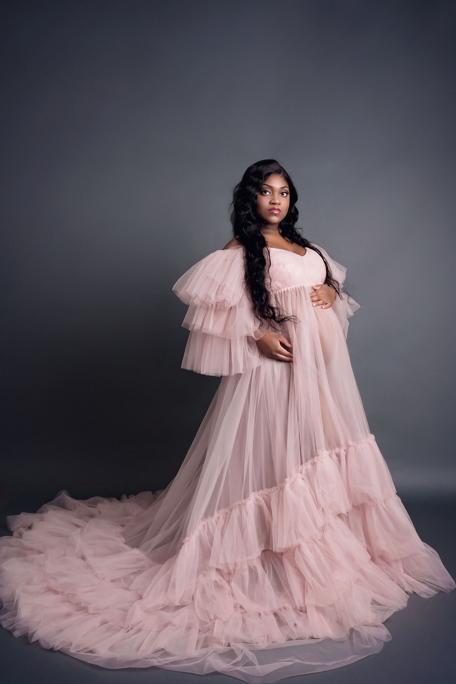 Dramatic maternity photo featuring a flowing pink tulle gown with tiered ruffles against a dark gray background.