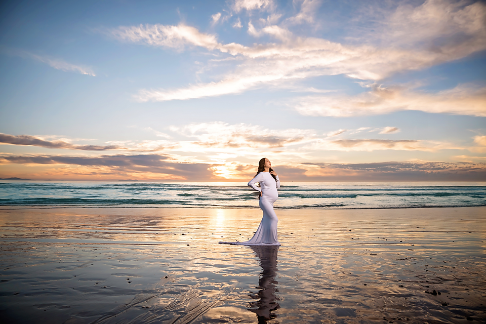 Pregnant woman in white dress standing on beach at sunset with golden sky and ocean reflections.