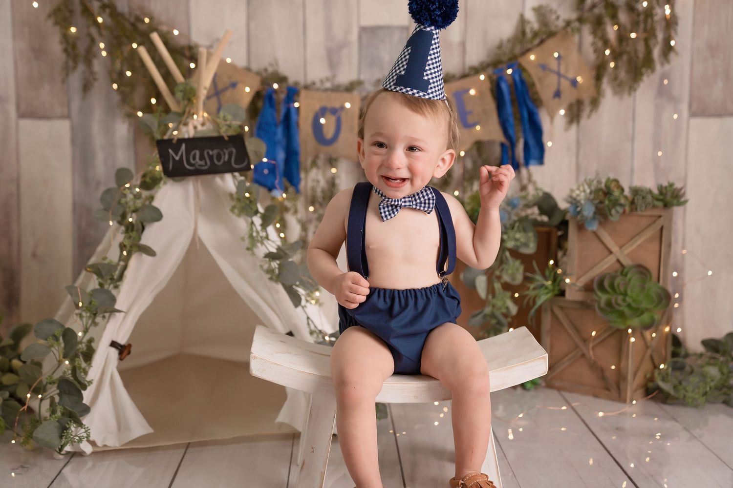 Baby in blue outfit sits among rustic woodland decorations with garland and twinkle lights. San Diego cake smash photographer