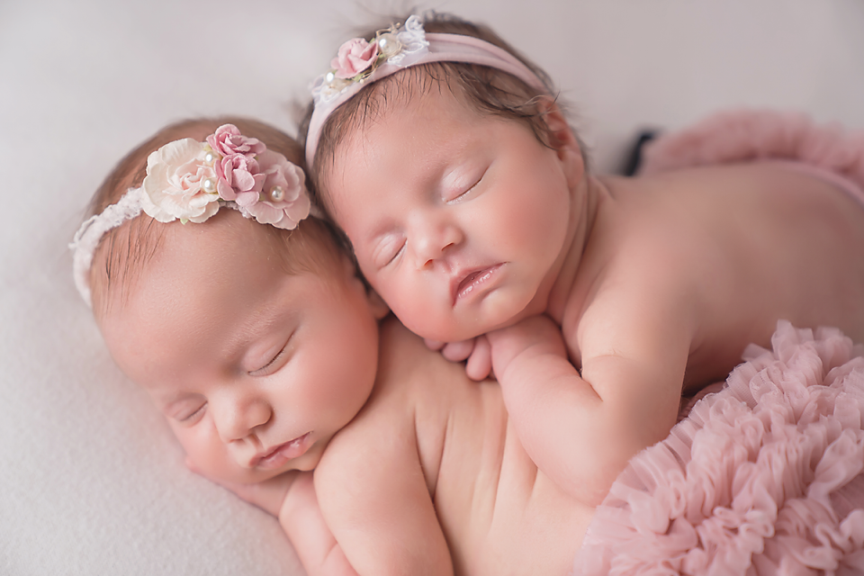 Two sleeping newborn twin baby girls wearing pink floral headbands, snuggled together in soft pink tutu skirts.