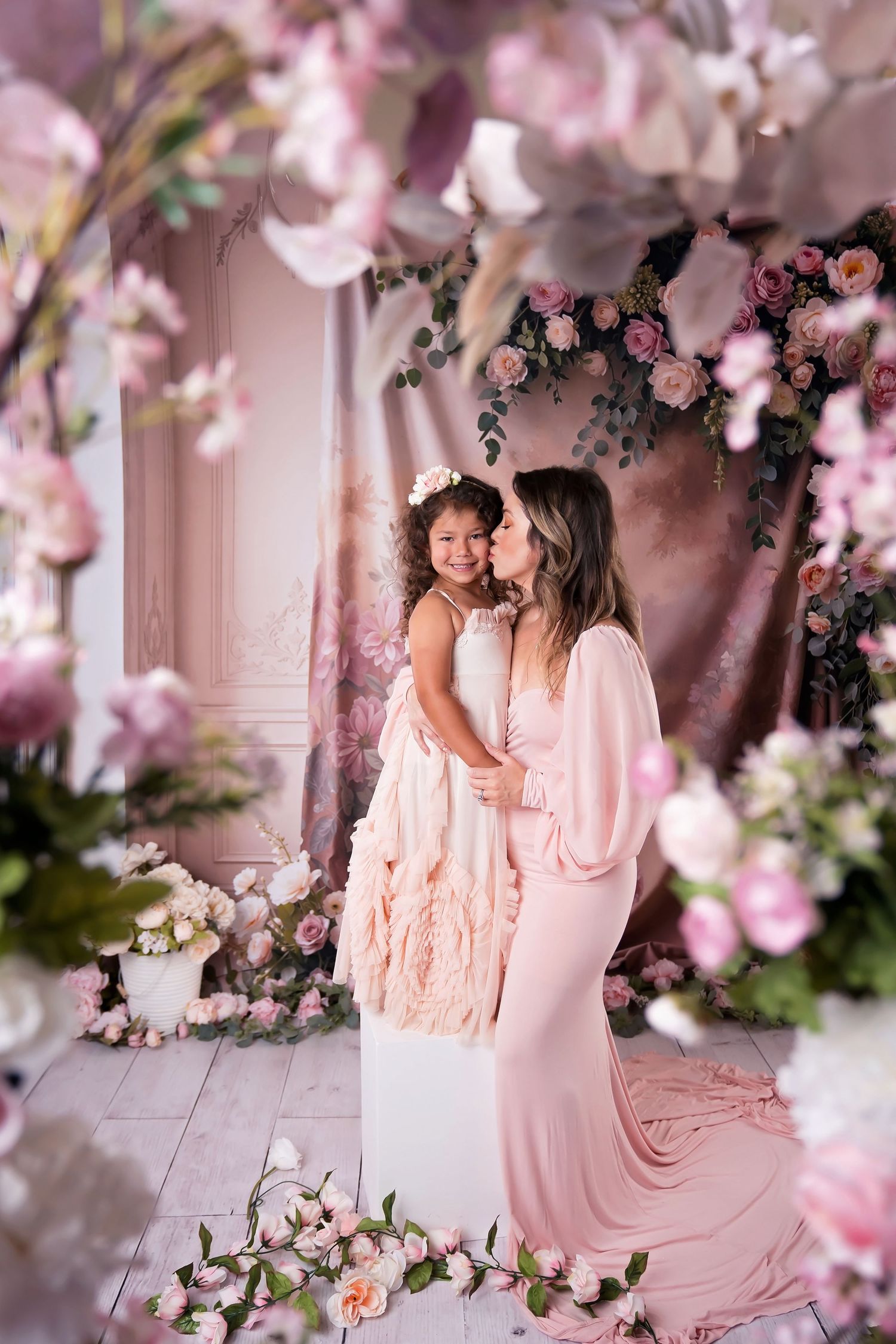 A mother in a pink gown kisses her young daughter surrounded by beautiful pink flowers on a mother's day photoshoot.