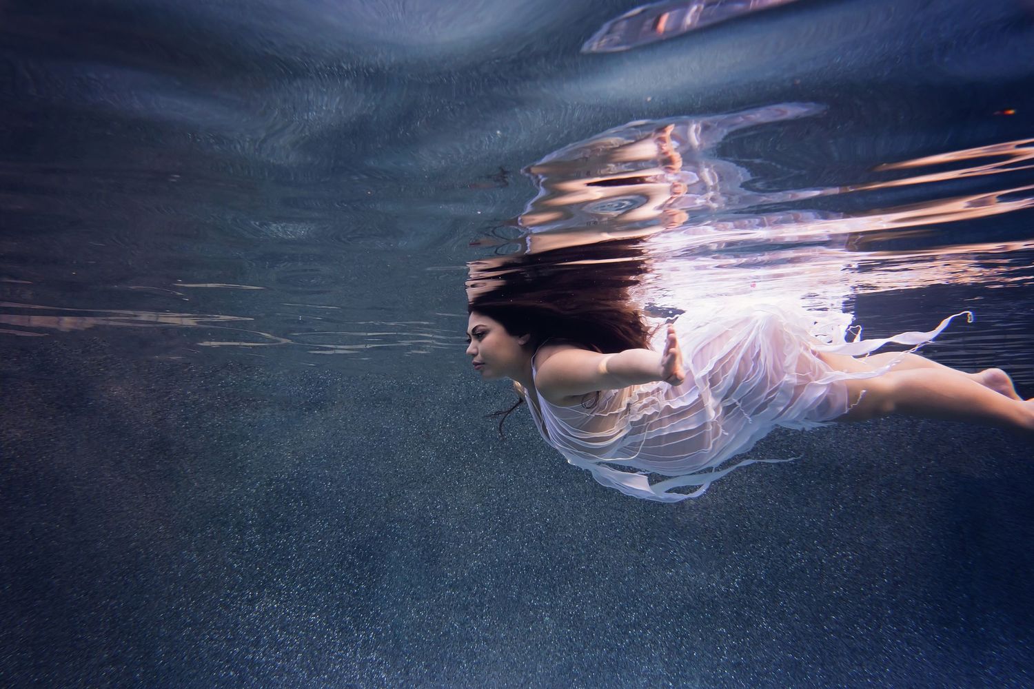 Artistic underwater maternity portrait with flowing fabric and glowing light trails in deep blue tones