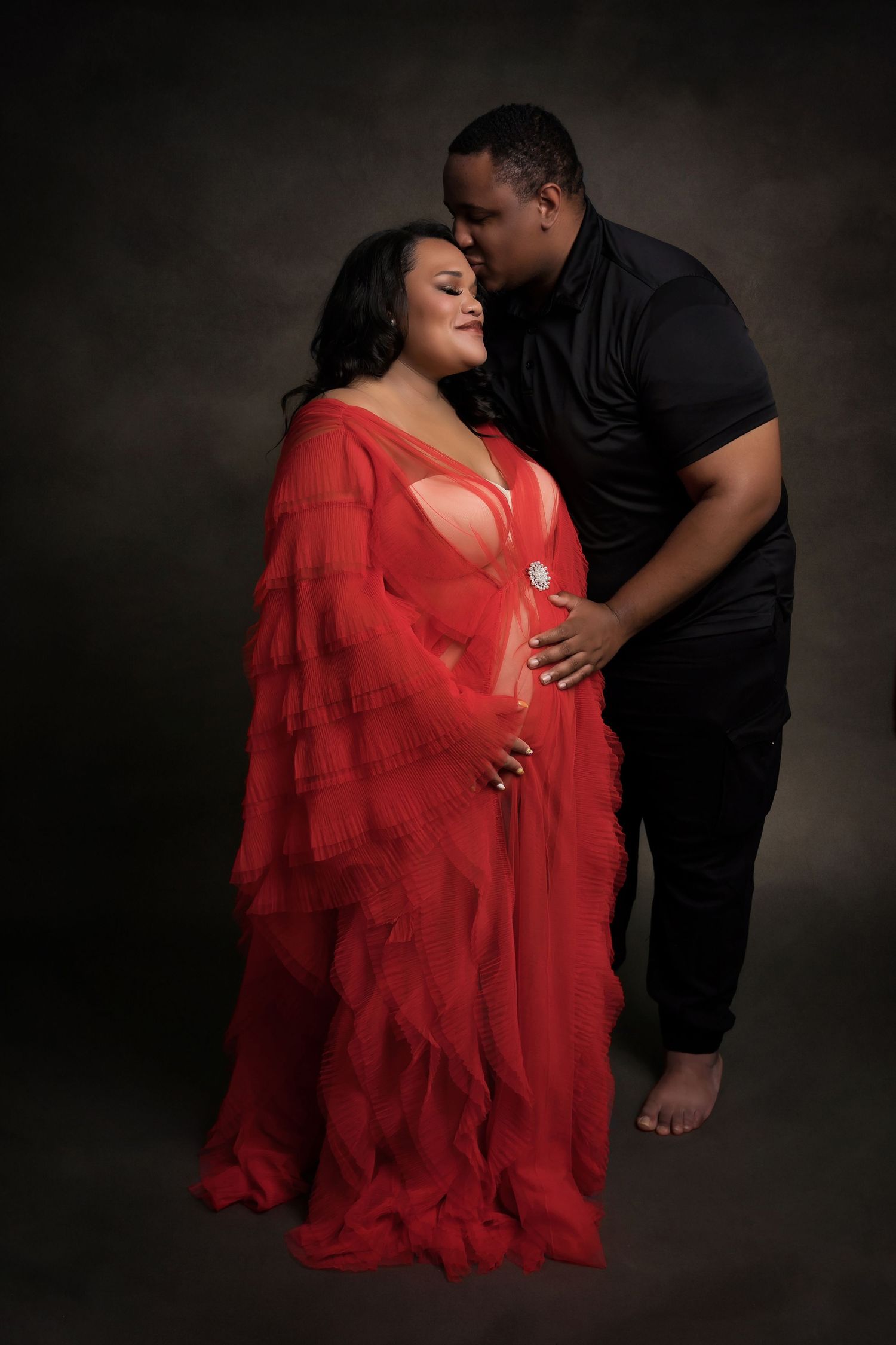 Intimate maternity portrait with couple in red and black attire against dark backdrop.