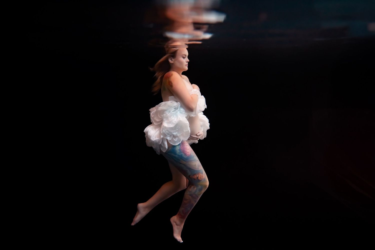 Pregnant woman in white gown floating underwater with soft movement against a dark backdrop — San Diego maternity session