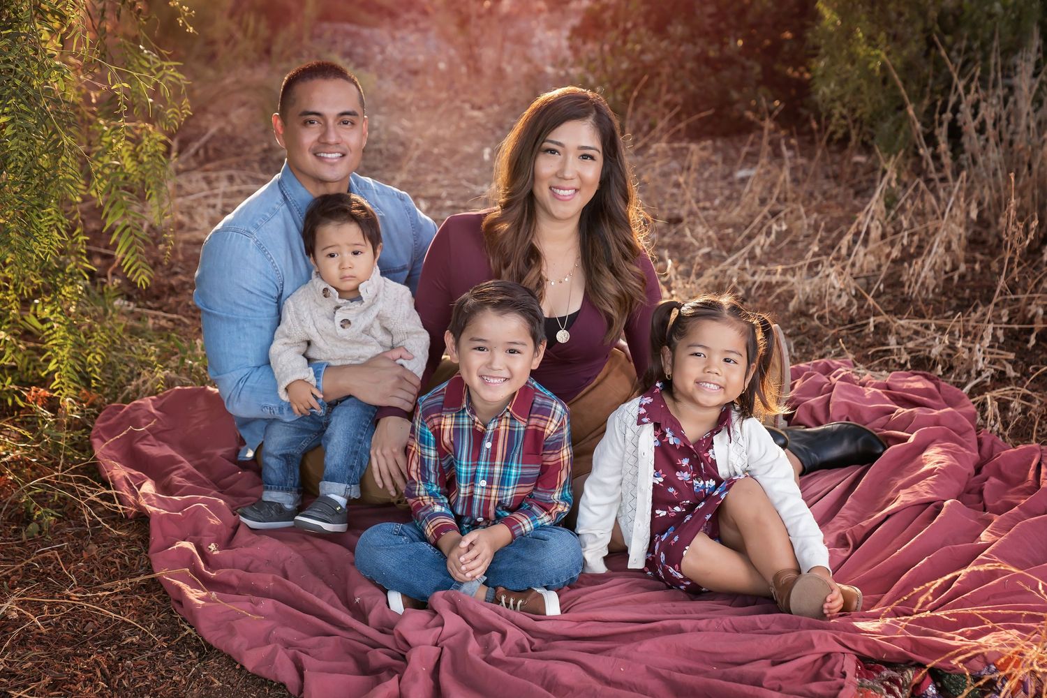 Family relaxing together on a burgundy blanket in a wooded outdoor setting during golden hour.