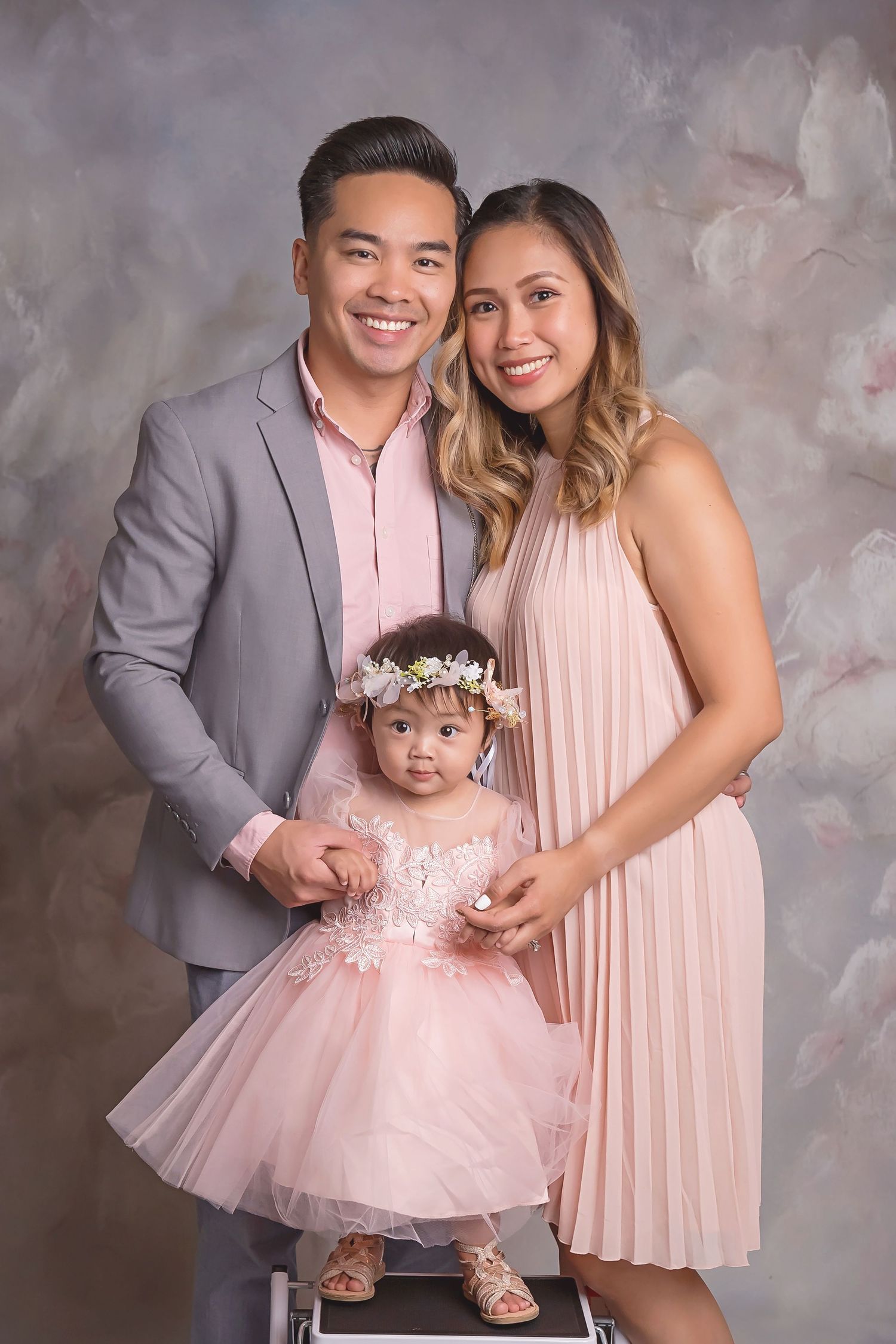 Parents holding their baby in coordinated pink and gray outfits against a mottled studio backdrop