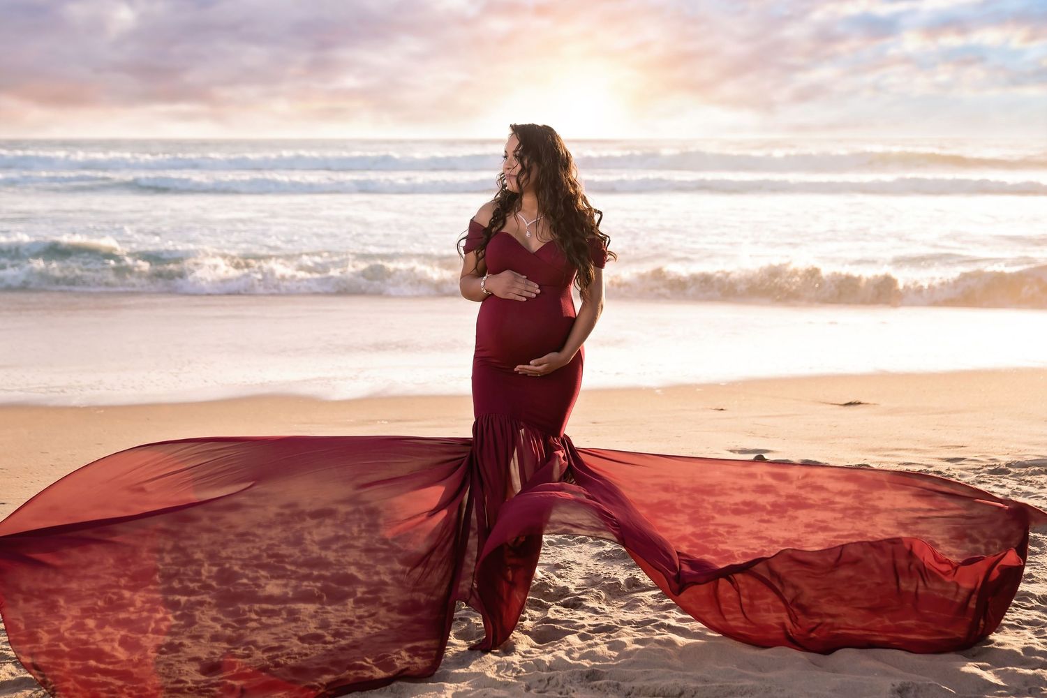 A flowing red dress catches the wind during a sunset maternity photoshoot on a beach. San Diego Maternity Photographer