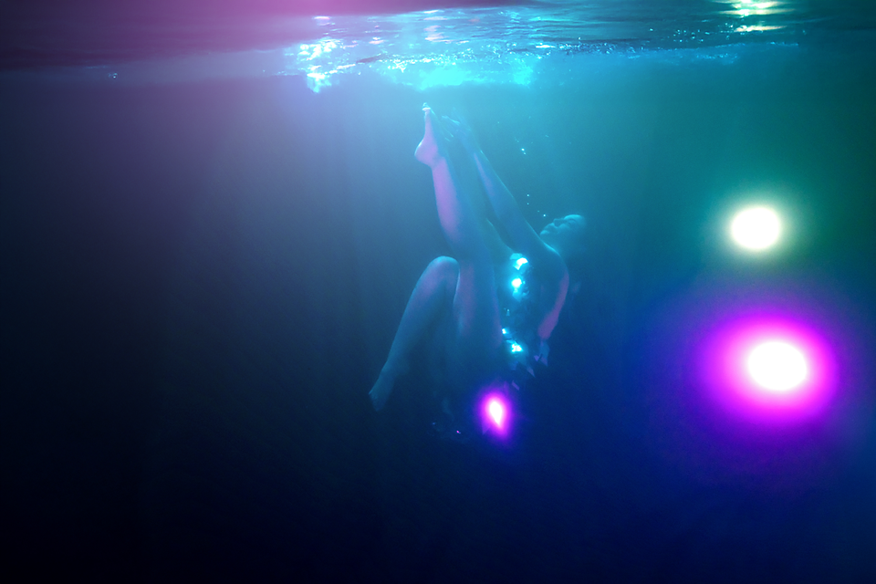 Person diving underwater surrounded by colorful glowing lights in dark blue water.