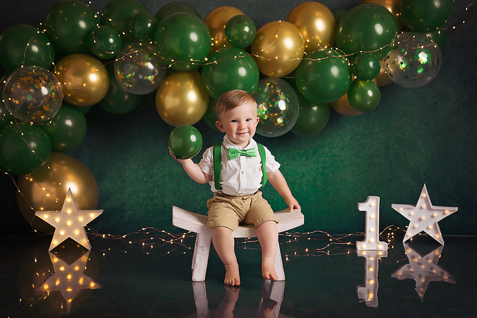 Smiling baby boy in suspenders sits among green and gold balloons celebrating his first birthday.