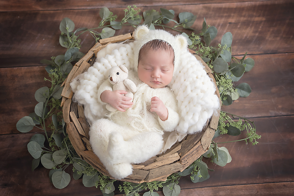 Newborn baby in white bear outfit sleeping in a rustic basket with green eucalyptus leaves and a teddy bear.