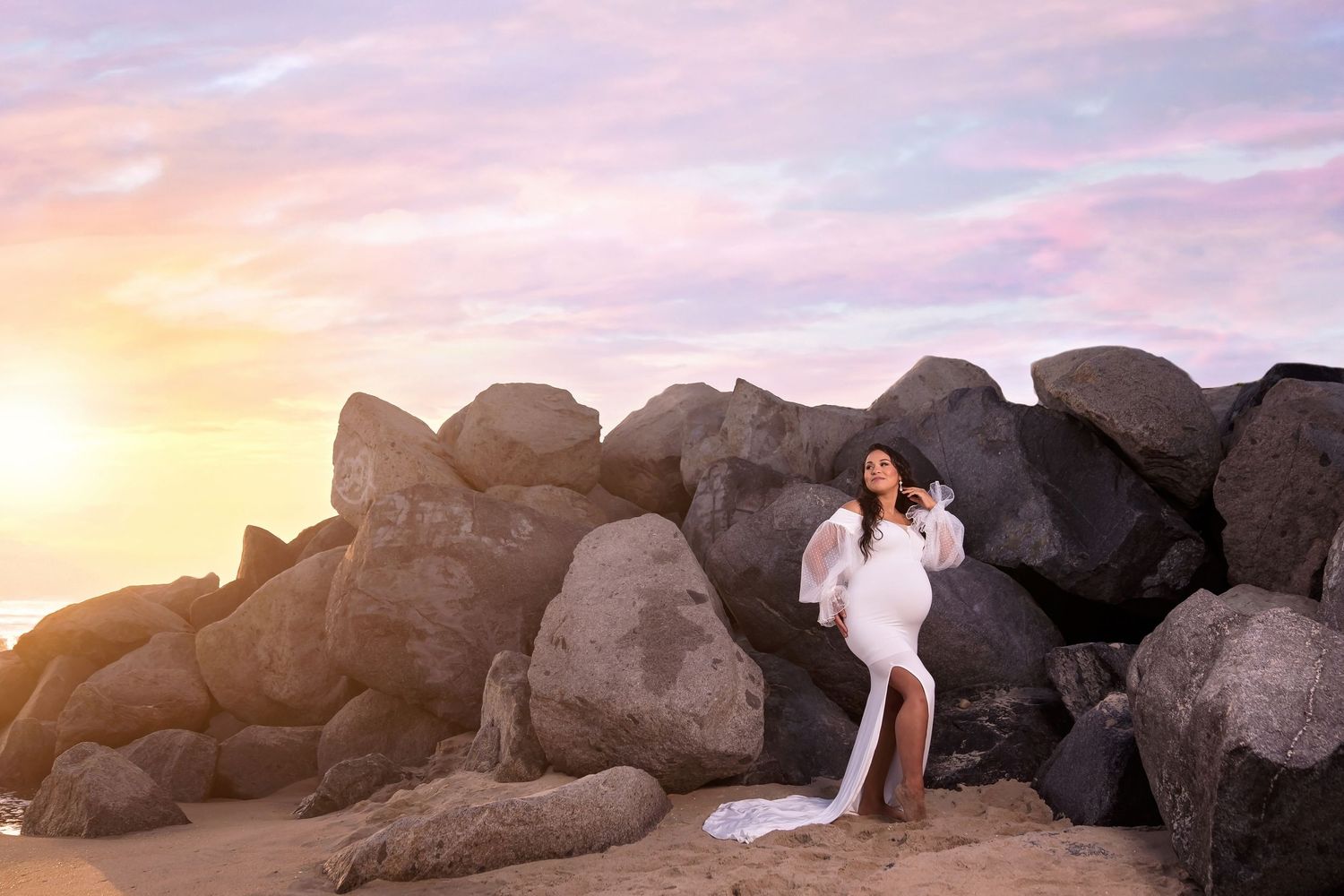maternity photo in white dress contrasts against dark coastal rocks during a golden sunset beach session. San Diego Maternity Photographer