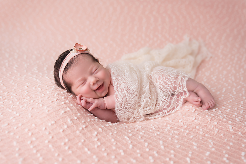 Smiling newborn baby girl swaddled in cream lace wrap with pink bow headband on pink textured blanket.
