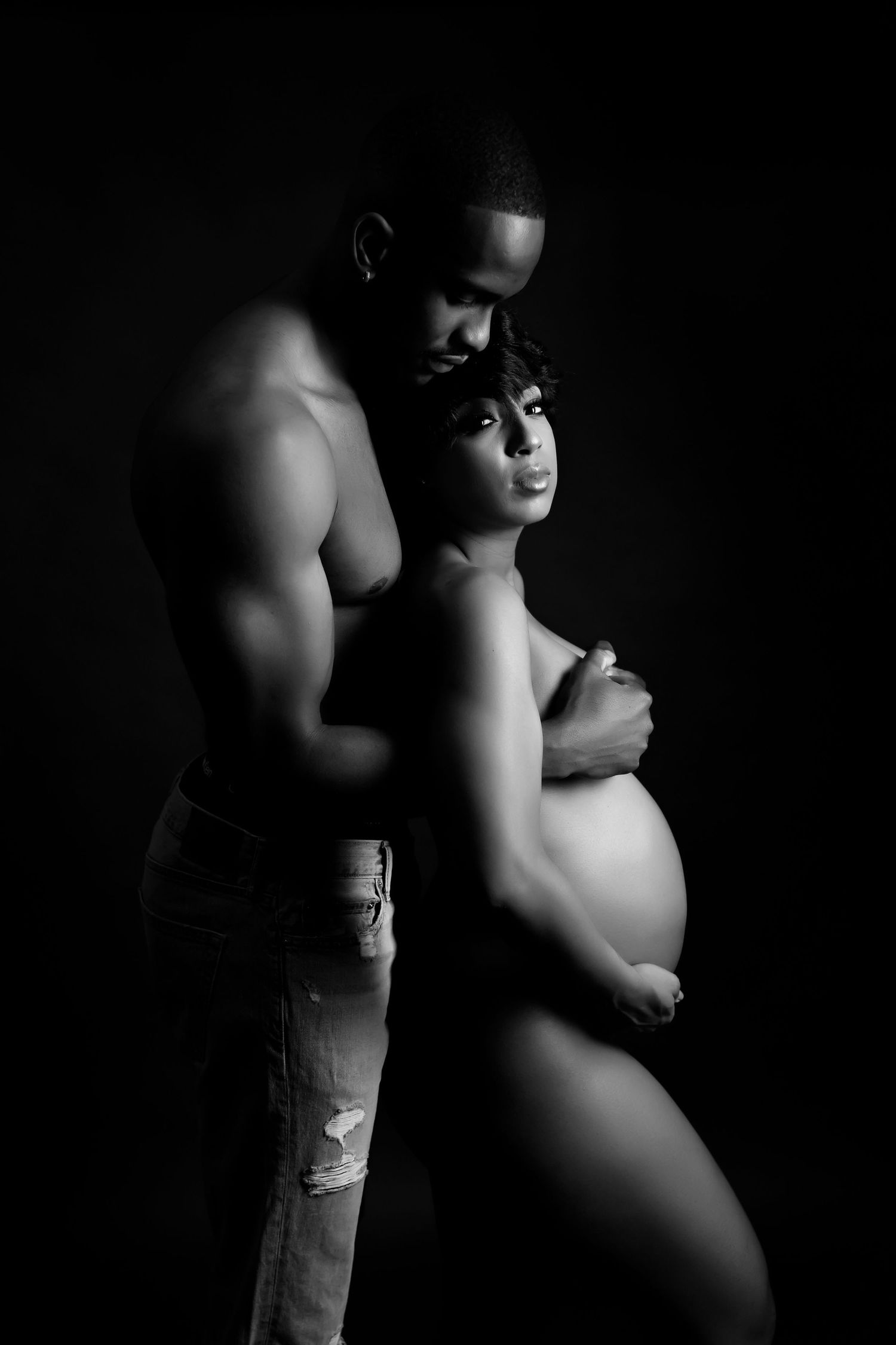 Dramatic black and white maternity portrait with couple against dark background.