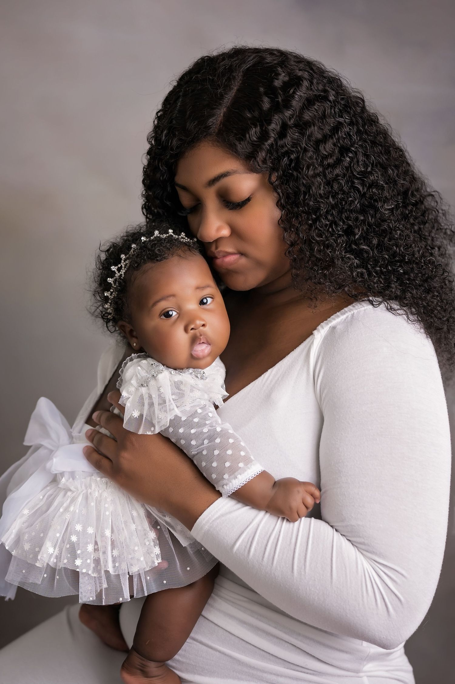 Mother and baby in matching white dresses sharing a tender moment against gray studio backdrop