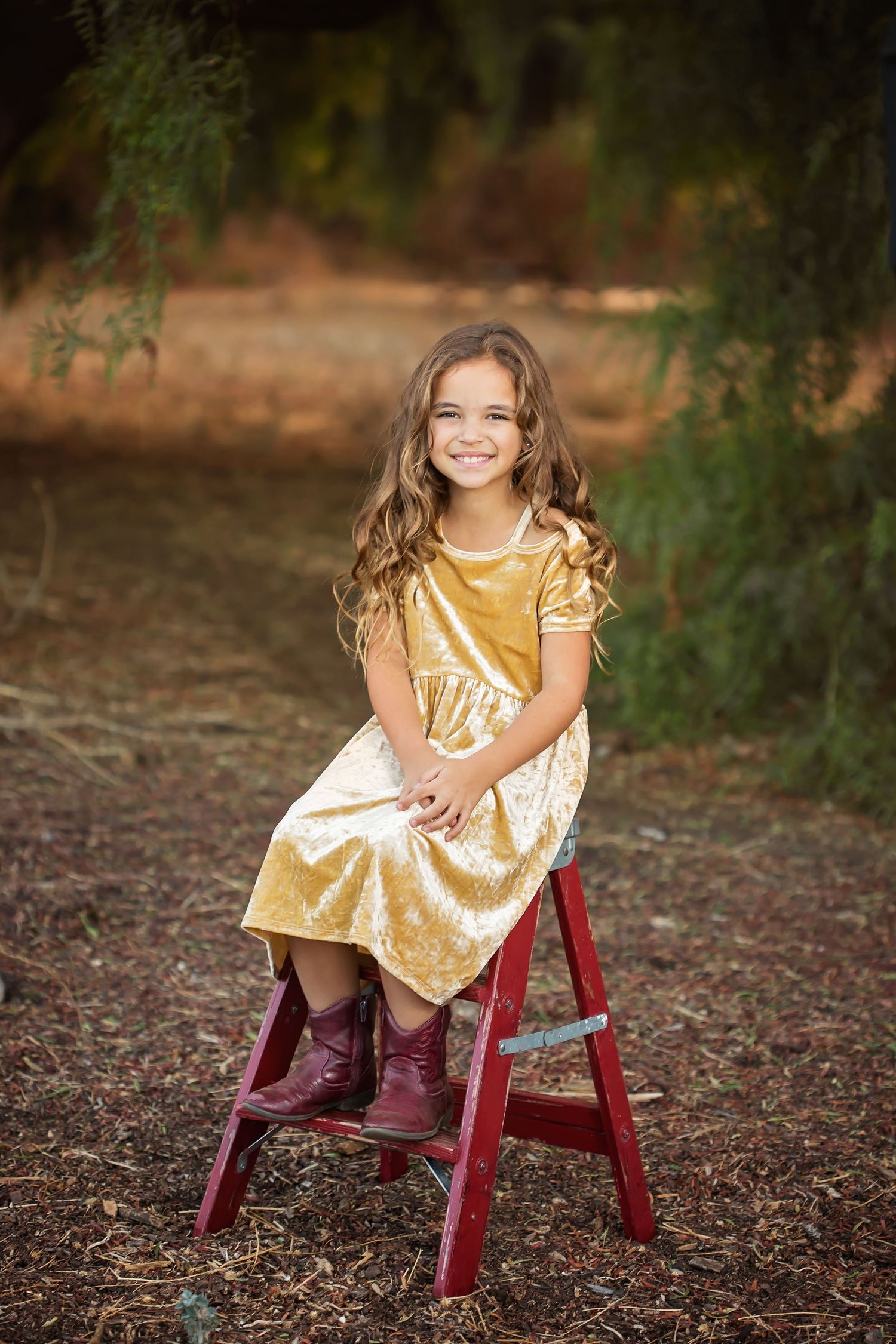 Child in a gold dress seated on a red wooden chair at golden hour in an outdoor setting