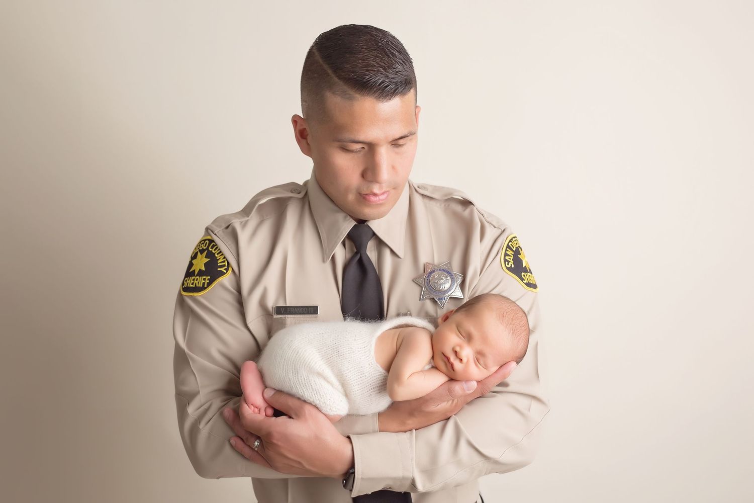 Police officer in tan uniform gently cradling newborn baby during portrait session.San Diego Newborn Photographer
