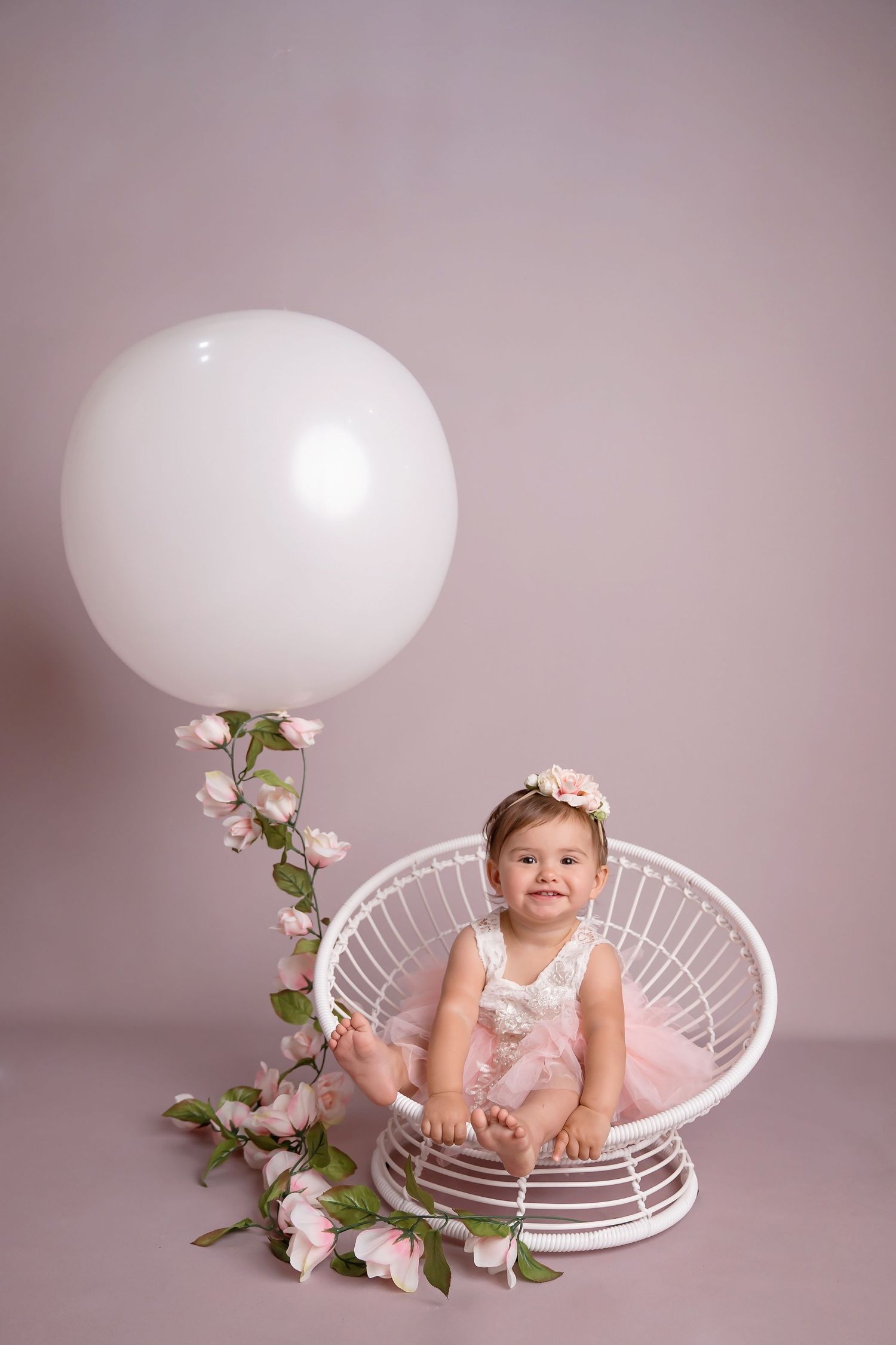 Baby sitting in white wire chair with floral decorations on pink background during portrait session.