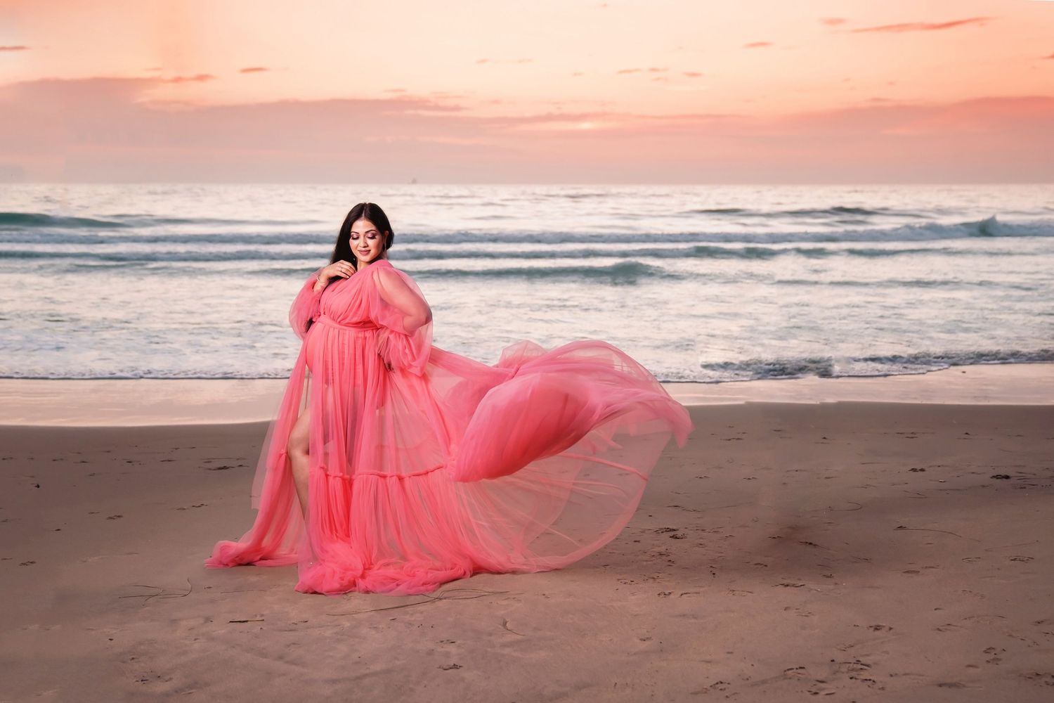 Dramatic sunset beach maternity photoshoot featuring flowing pink dress against coastal backdrop. San Diego Maternity Photographer