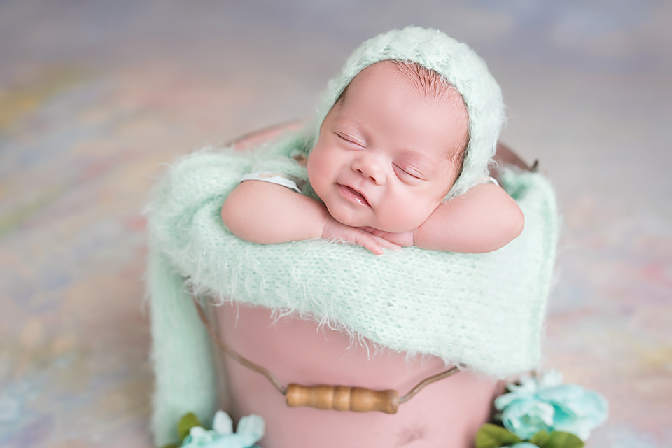 Sleeping newborn baby in mint green bonnet and wrap posed in a pink bucket with floral decorations.